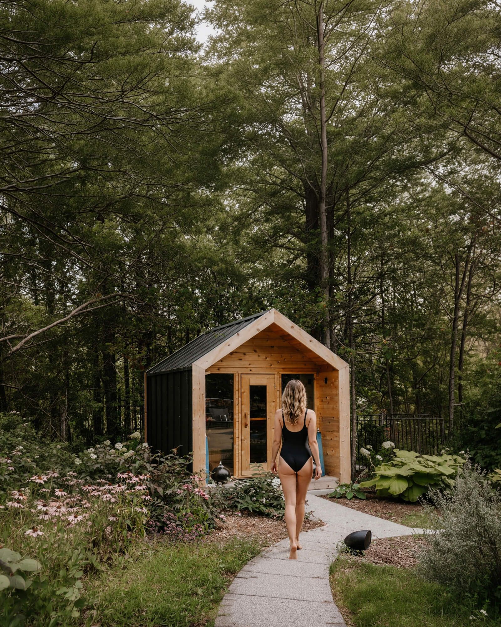 Woman in a black swimsuit walking along a stone path toward a Canadian Red Cedar cabin sauna nestled among tall trees and wildflowers