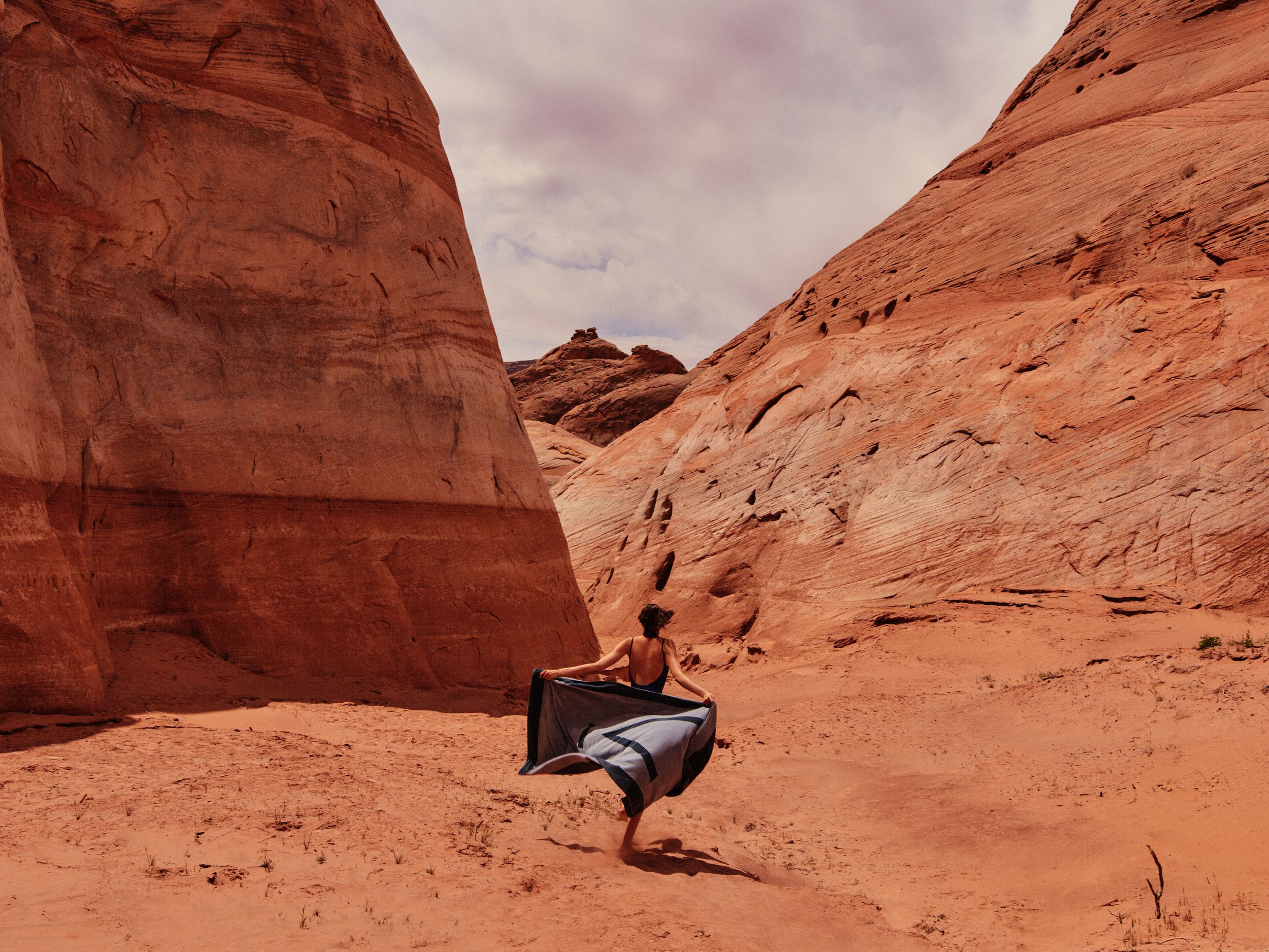 A model running through the red sands of the Utah desert holding an Aman towel behind her, towards the intersection of the feet of two rock formations. Photography by Chris Colls, Art Direction by Colville-Walker.