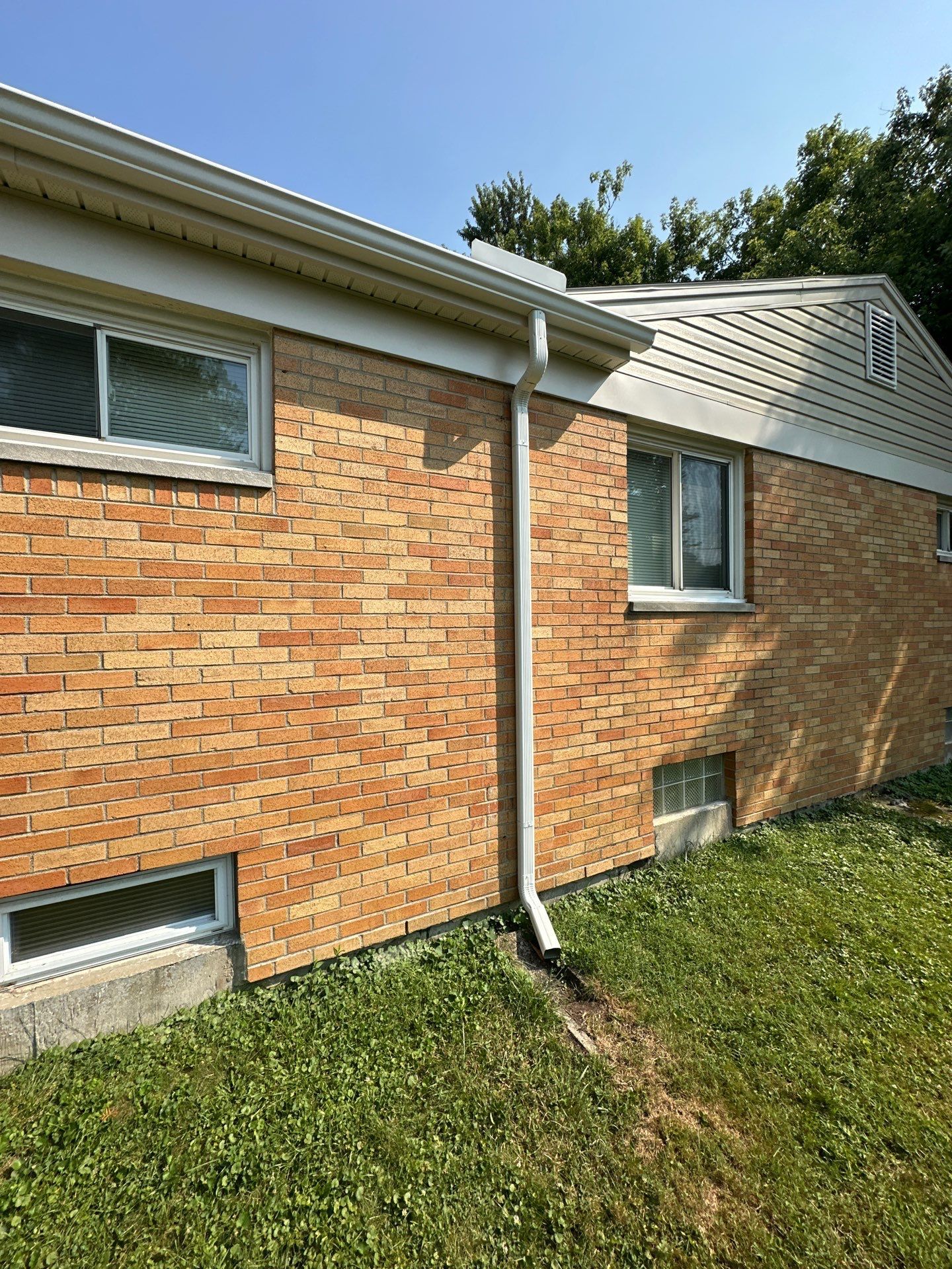 A new white seamless gutter running along the eave and around the corner of a brick home, with a downspout carrying water to the ground, in Dayton, Ohio.