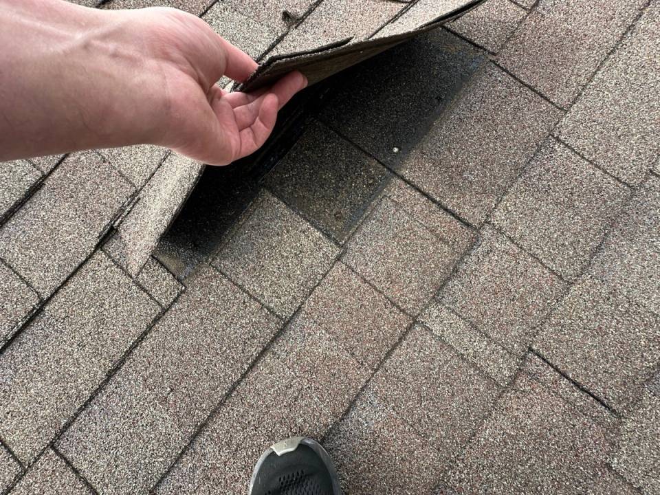 A hand lifting a shingle tab during a roof inspection.