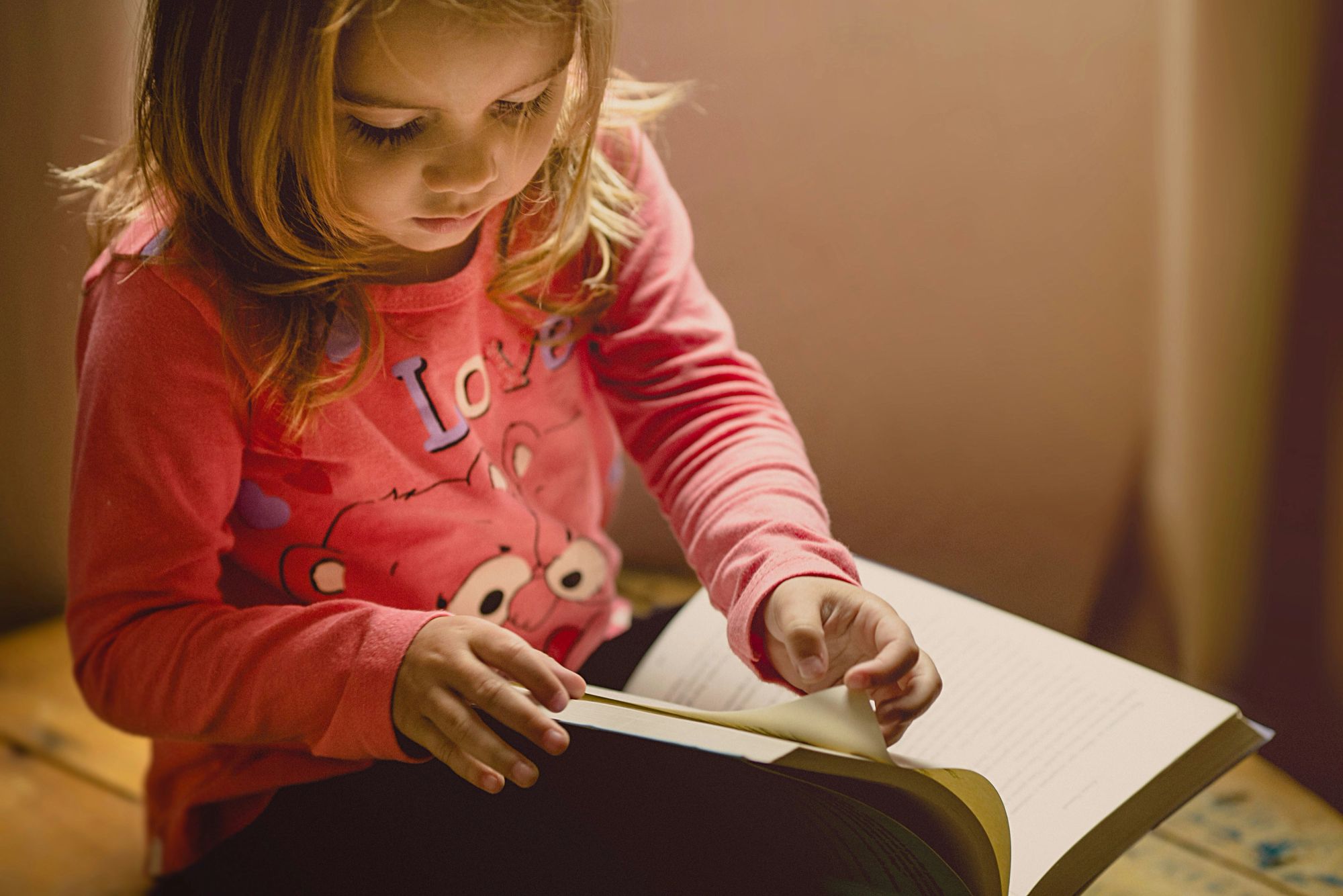 A girl reaching out to turn the page of an open book on her lap.