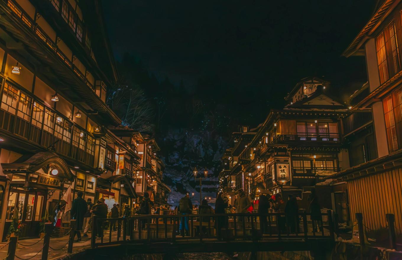 Ginzan Onsen at night: lit wooden ryokan on both banks of the river, linked by small footbridges