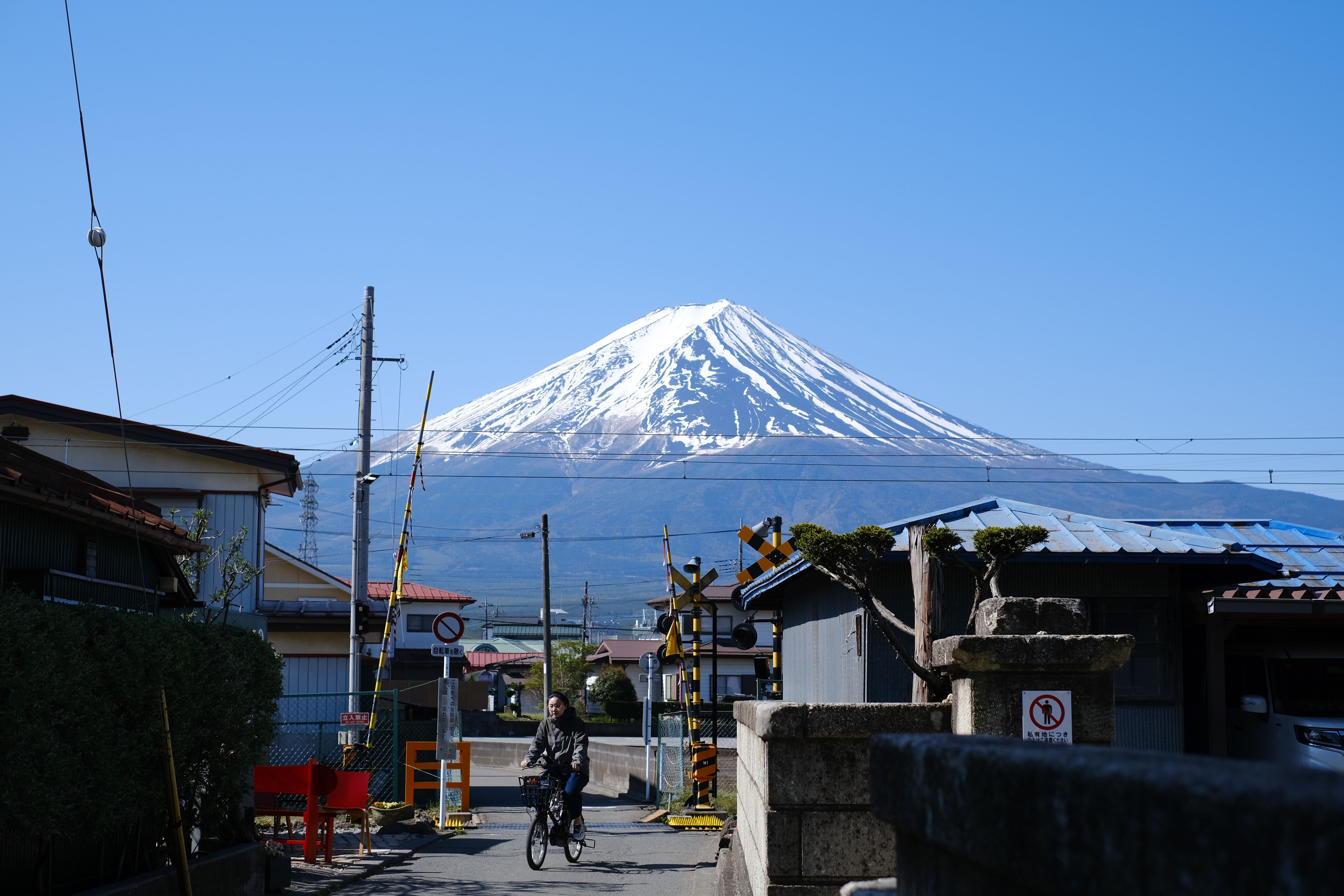 A cyclist passing a railway crossing with snow-capped Mount Fuji rising under a clear blue sky in Japan