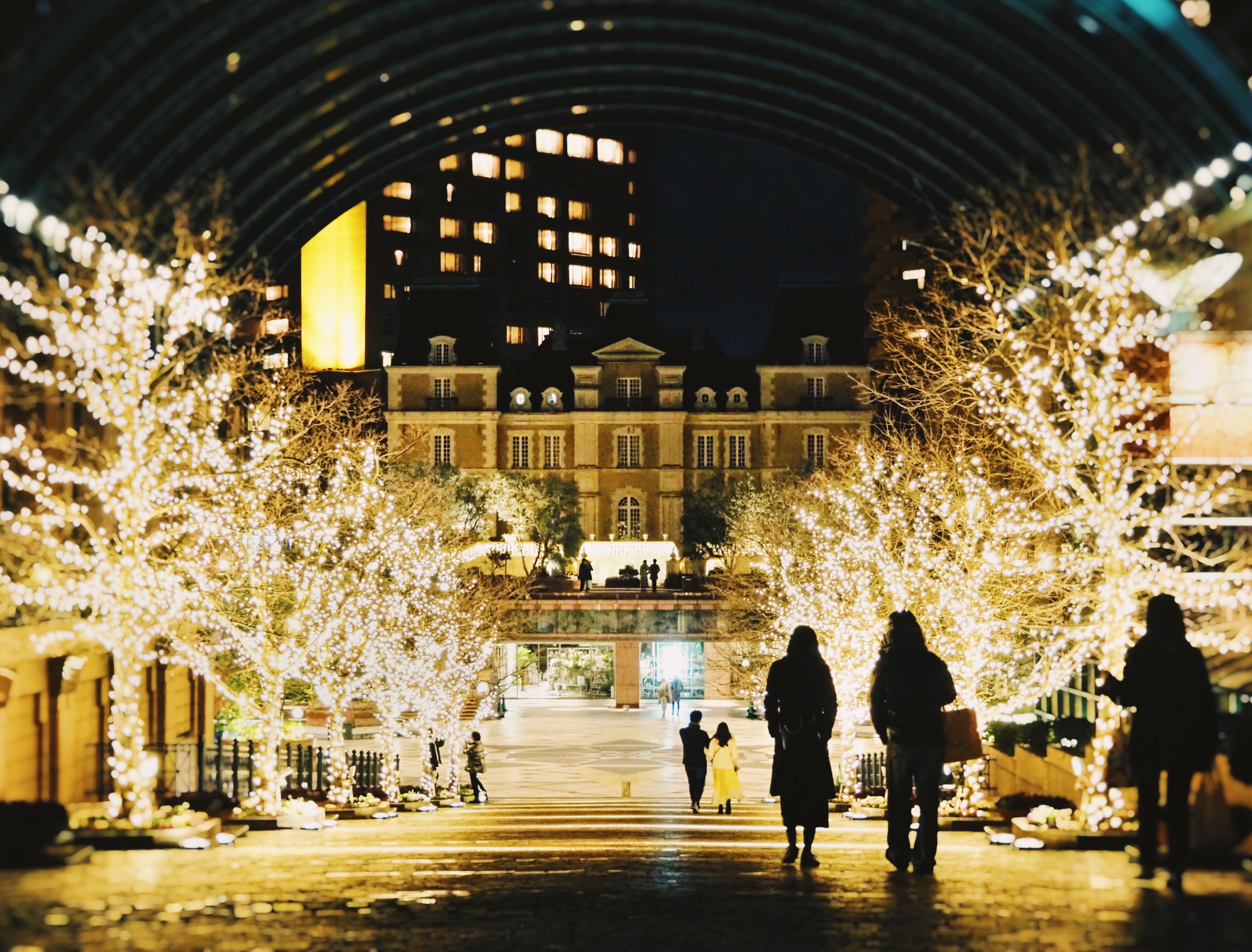 Trees wrapped in warm white lights under the glass arcade at Yebisu Garden Place, Tokyo, with people walking toward the chateau-style building at night
