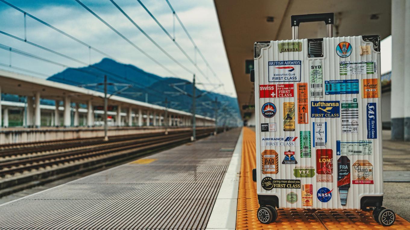 A white suitcase with travel stickers stands alone on an empty Japanese railway platform with mountains behind