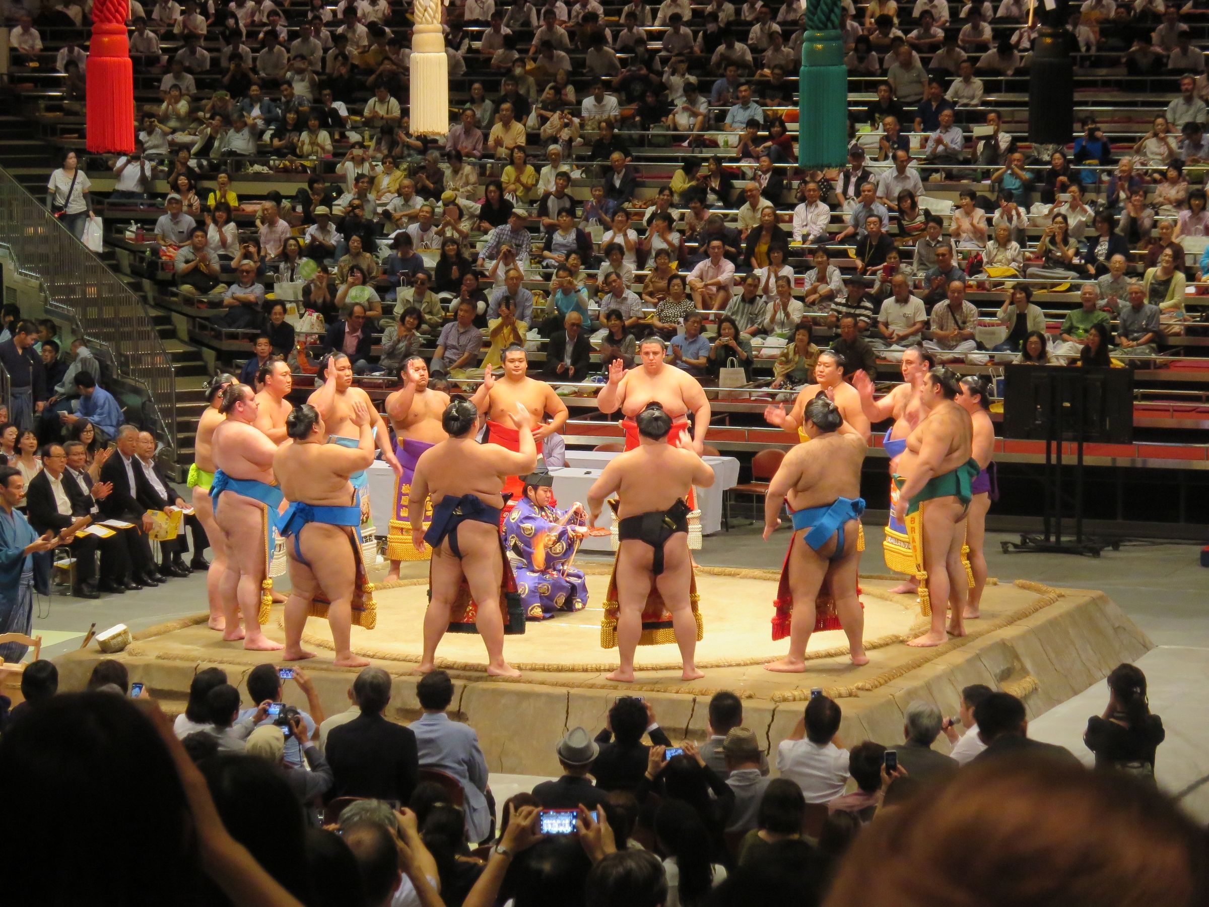 Sumo wrestlers gathered around the dohyo ring during a grand tournament in a packed Tokyo arena