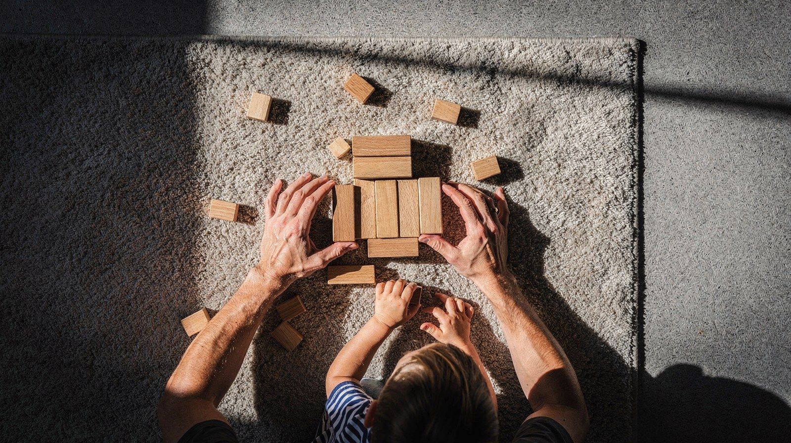 Un adulto e un bambino costruiscono con blocchi di legno su un tappeto, visti dall’alto.