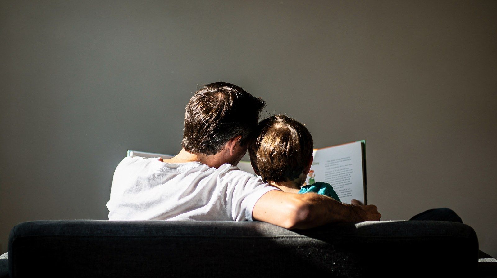 Un genitore tiene un libro illustrato mentre un bambino si avvicina, visti di spalle.