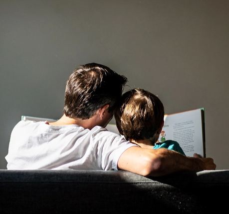 Un genitore tiene un libro illustrato mentre un bambino si avvicina, visti di spalle.