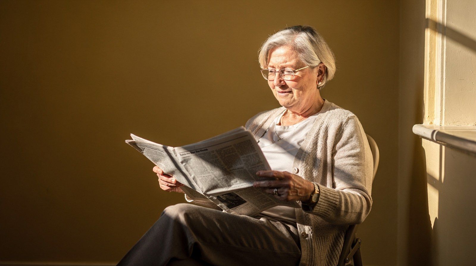 Una donna sui settant’anni legge il giornale accanto a una finestra soleggiata.