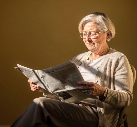 Una donna sui settant’anni legge il giornale accanto a una finestra soleggiata.