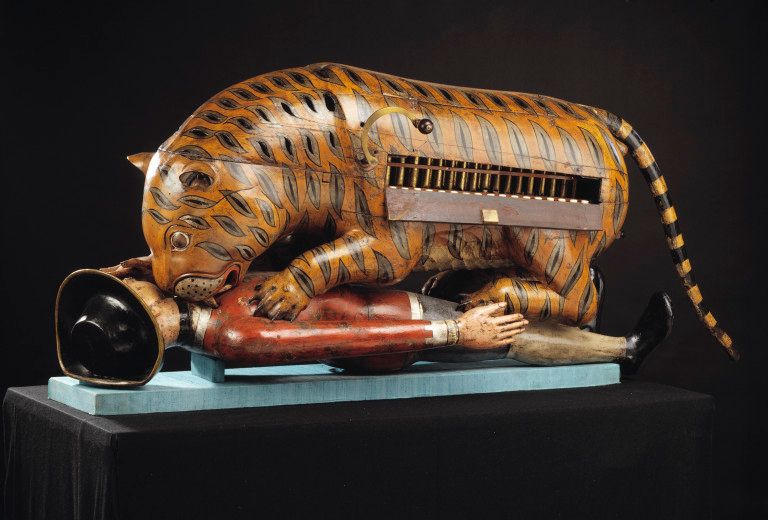 A painted wooden tiger crouching over a European figure, with the organ exposed in its side.