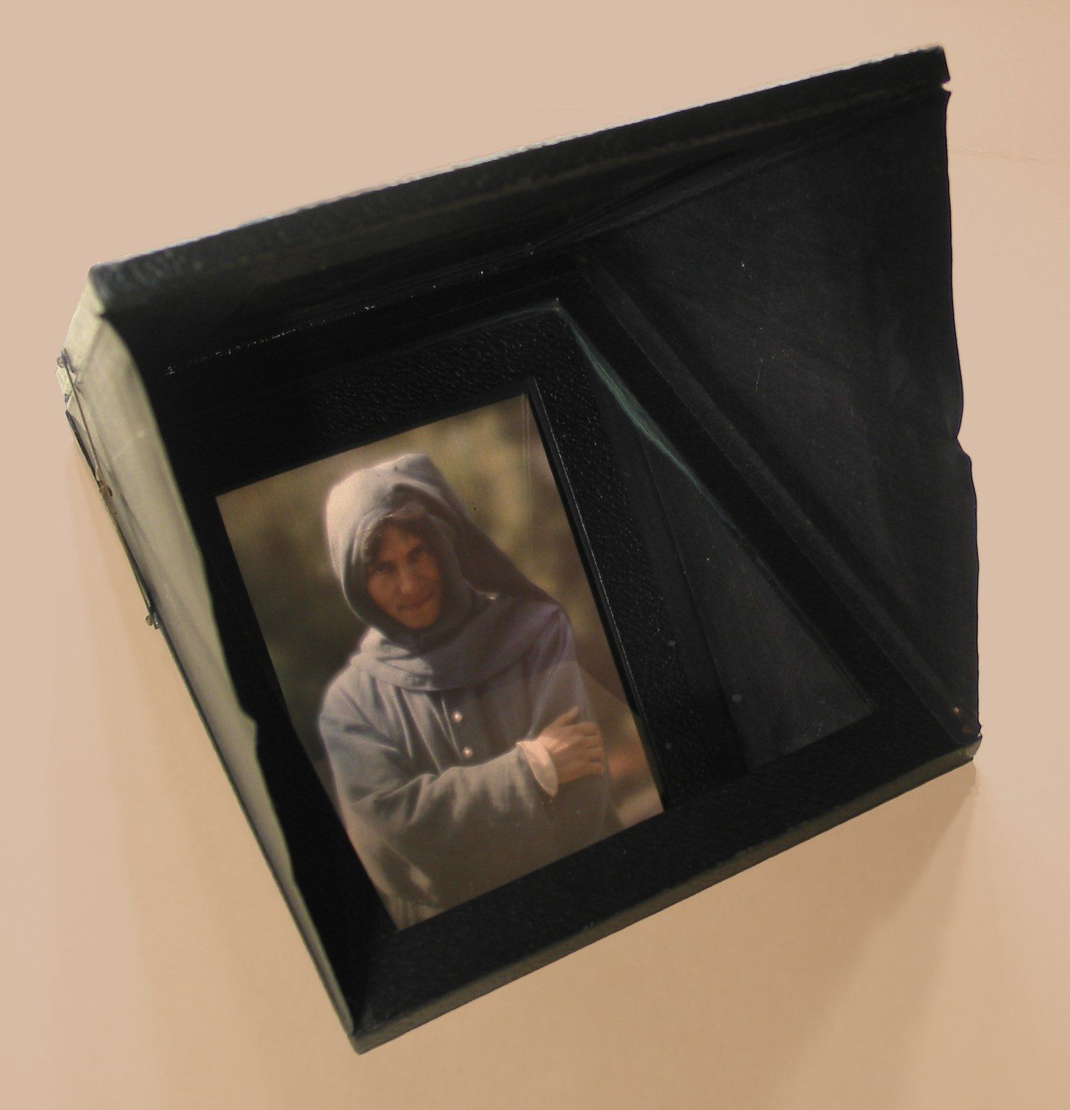 A folding black diascope with a colour portrait of a hooded man visible in its mirror.