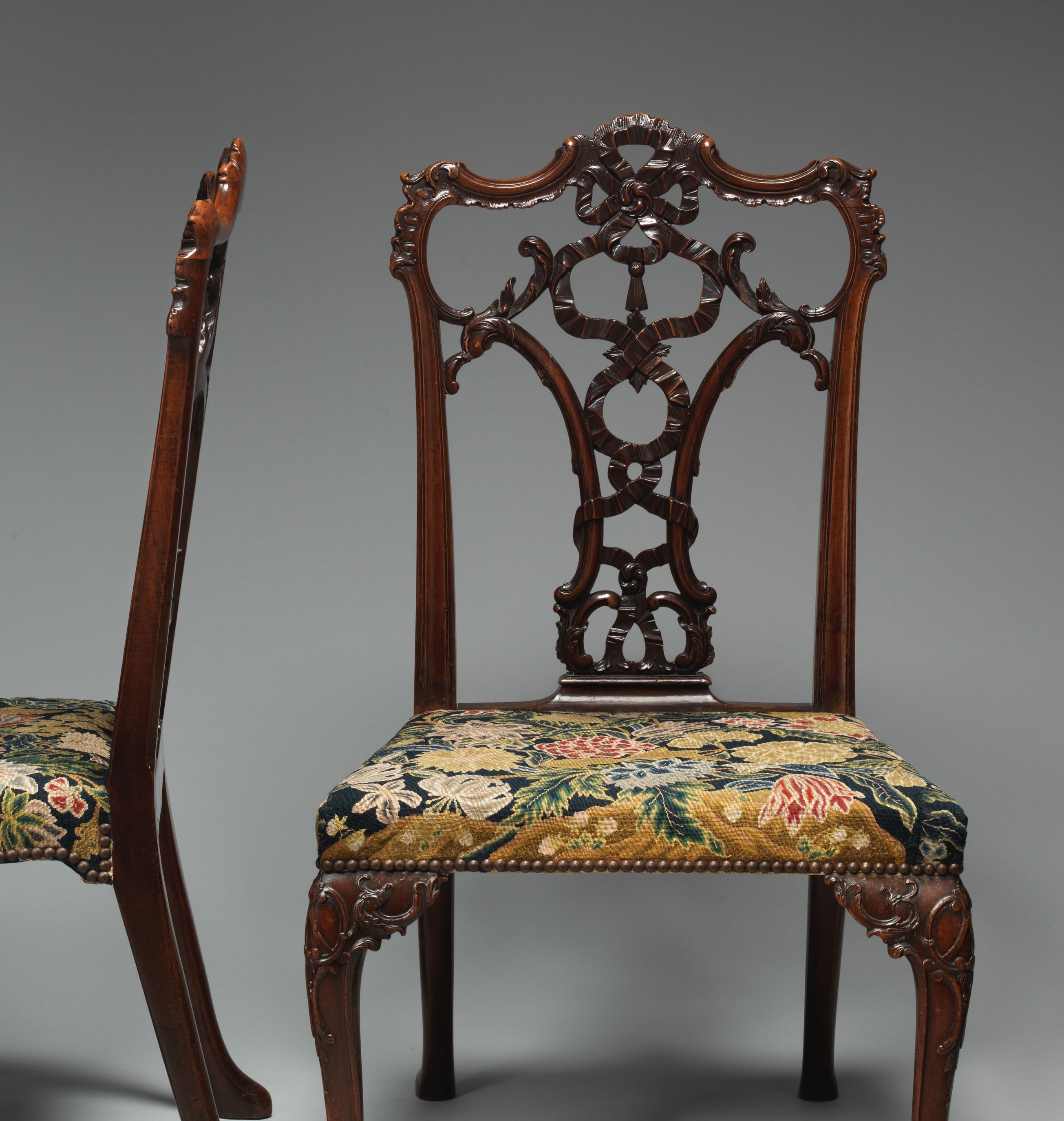 A mahogany chair with a carved ribbon back and flowered embroidered seat, beside part of its companion