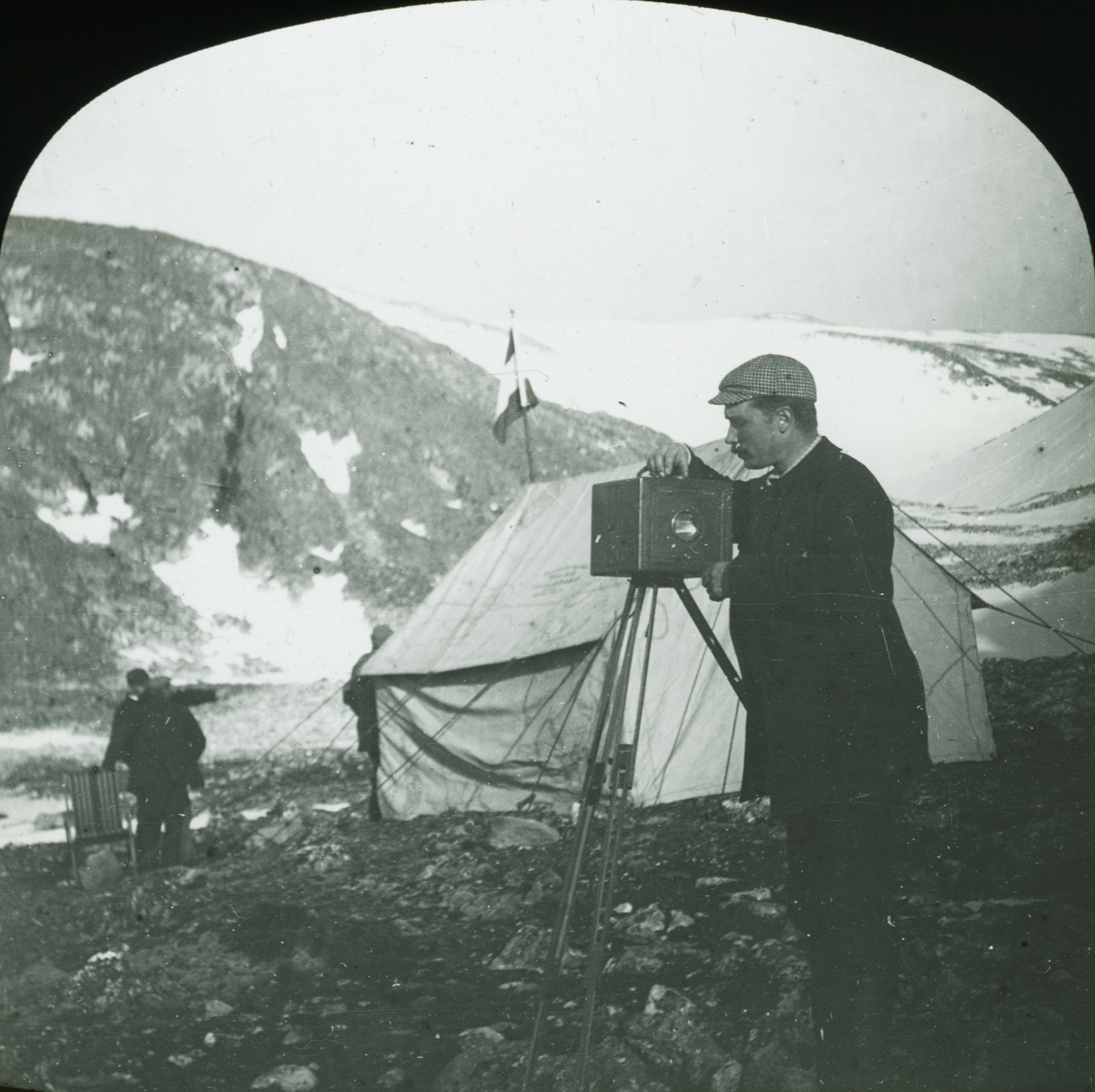 Nils Strindberg adjusting a wooden camera on a tripod, with a tent and mountains behind him