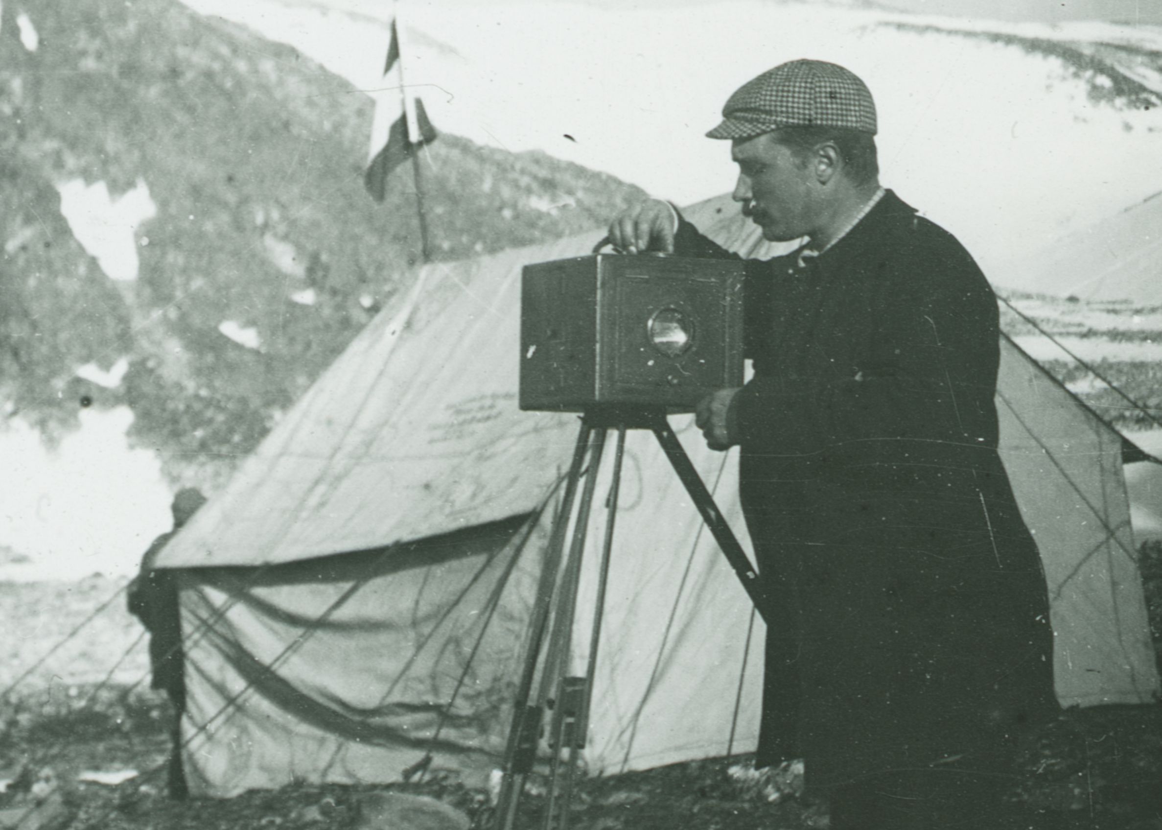 Nils Strindberg adjusting a wooden camera on a tripod, with a tent and mountains behind him