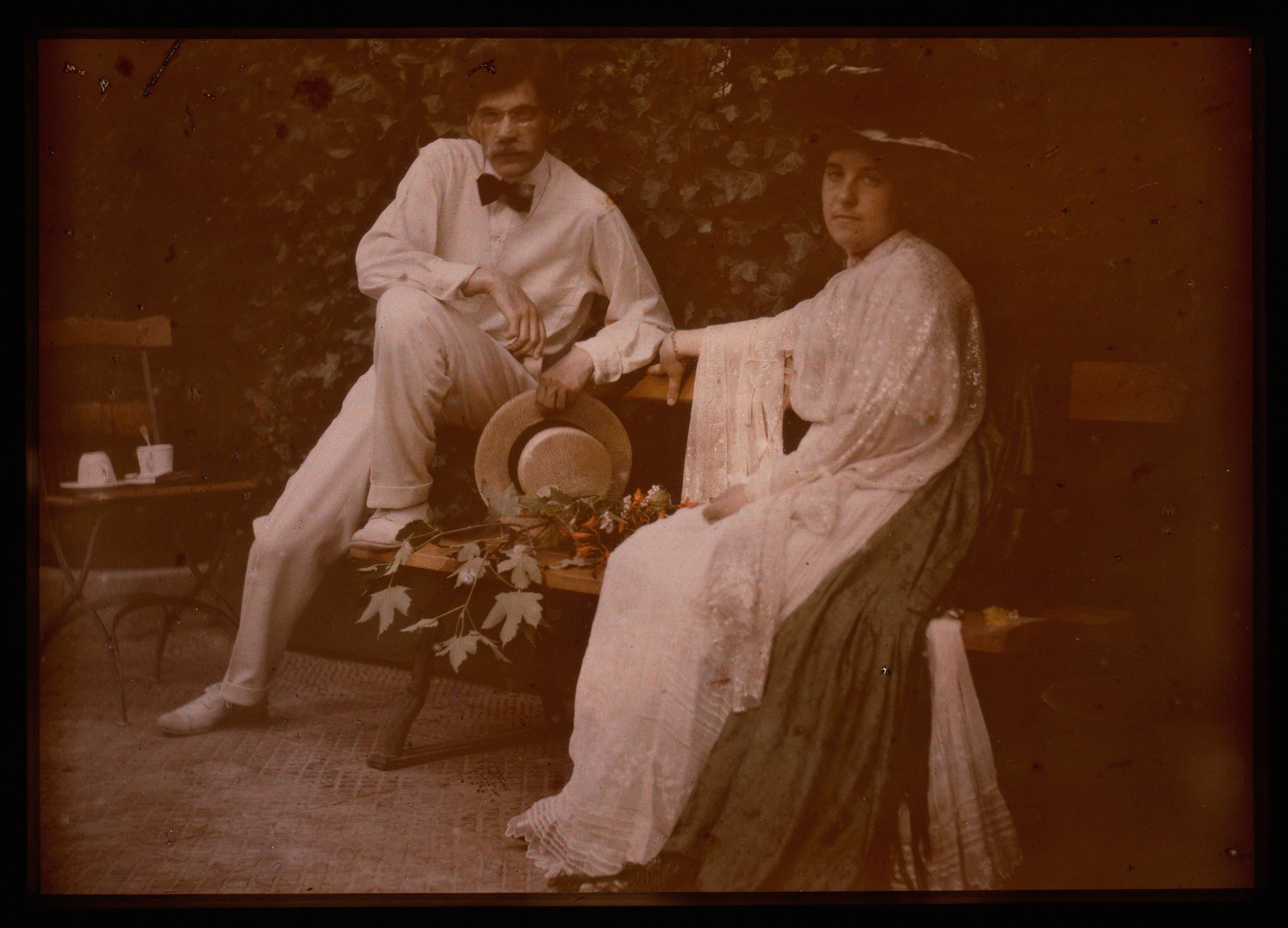 A man and woman in pale clothes sit among ivy, with flowers and a straw hat between them.