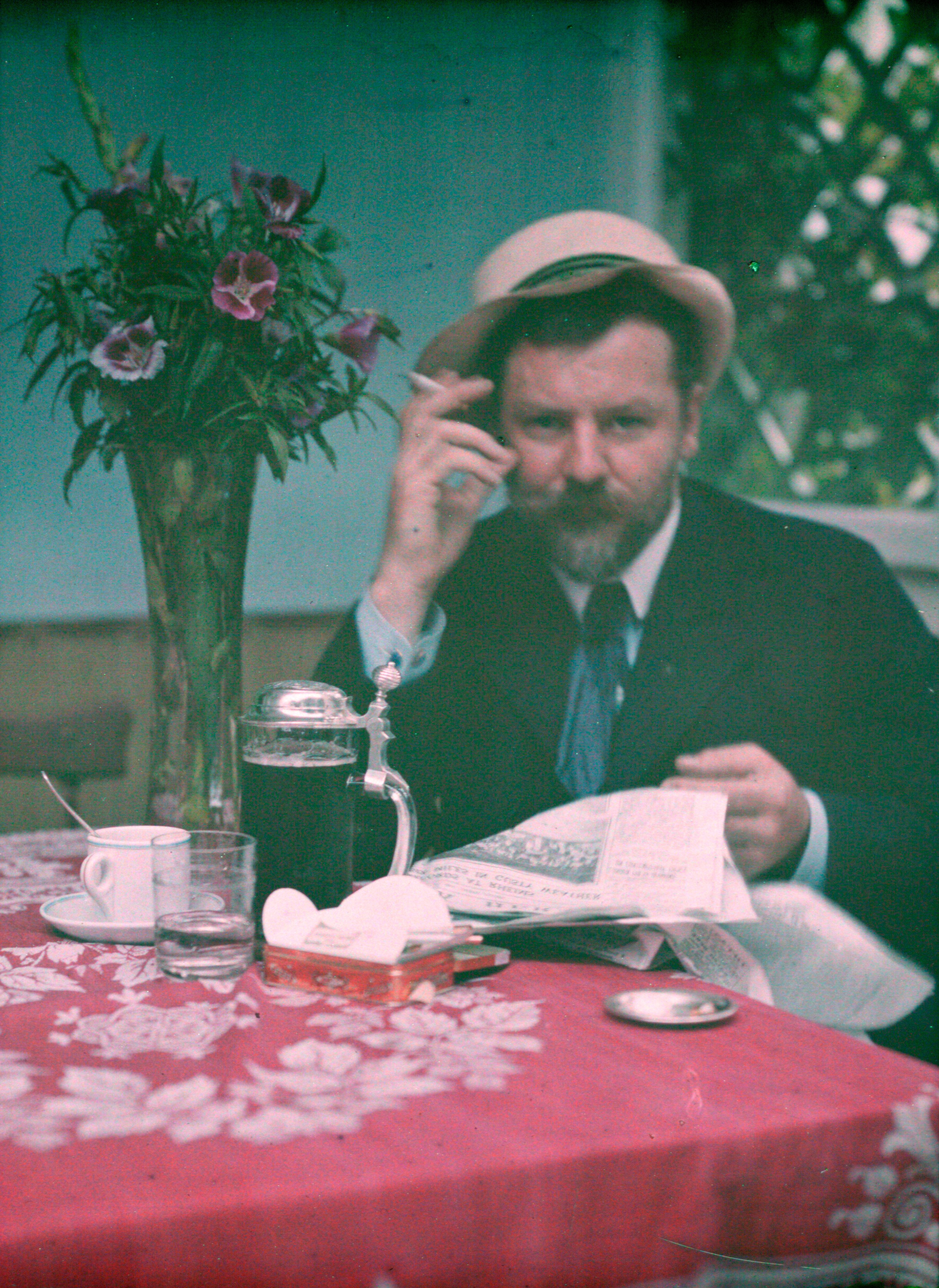 Frank Eugene sits at a table with a newspaper, cups, a beer stein and a vase of flowers; a red patterned cloth fills the foreground.