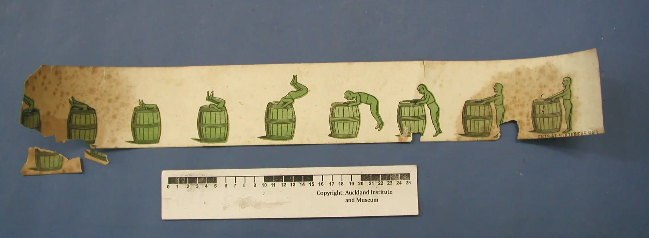A damaged paper strip showing a green figure bending into and emerging from a barrel, with detached fragments below