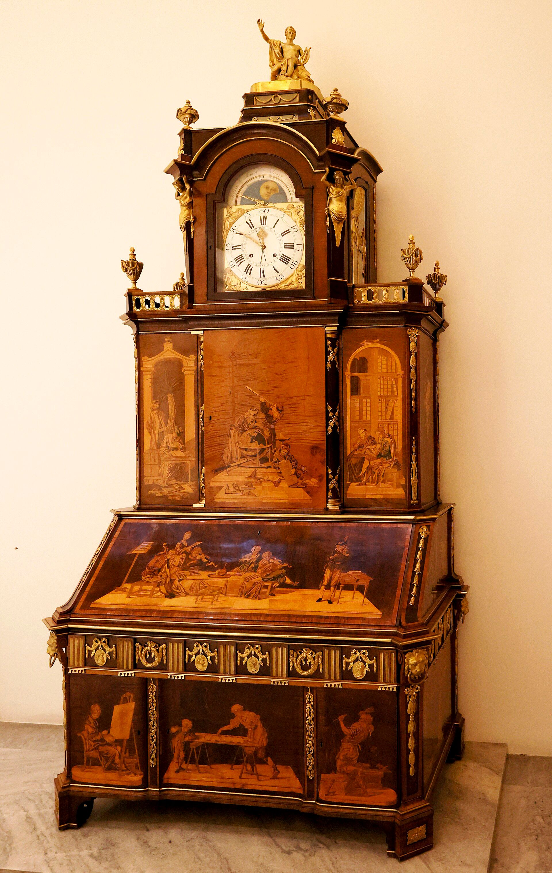 A tall marquetry cabinet with a writing flap, clock and gilded Apollo on its upper dome.