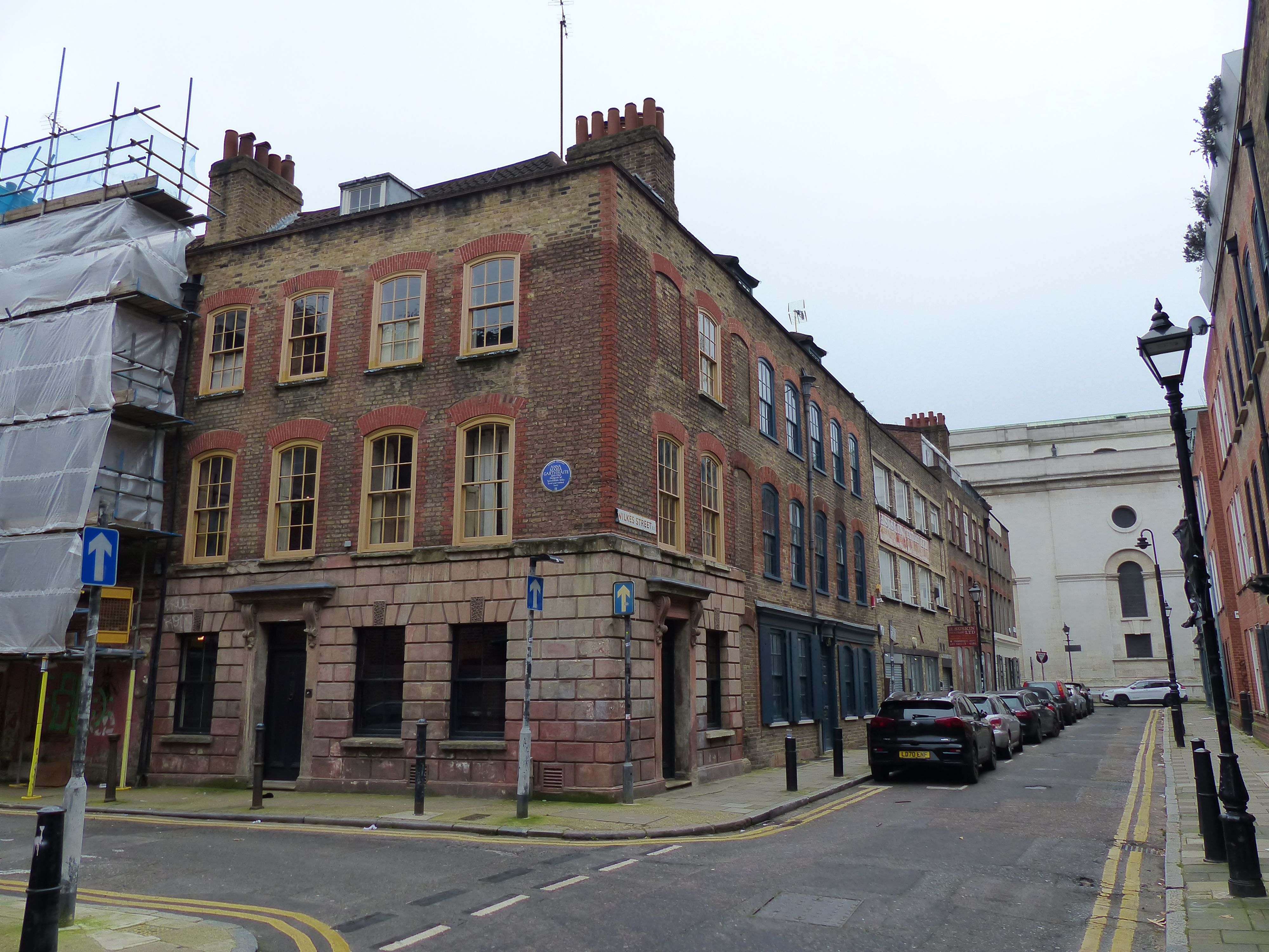 A brick corner house with yellow window frames and a blue plaque, on a narrow London street