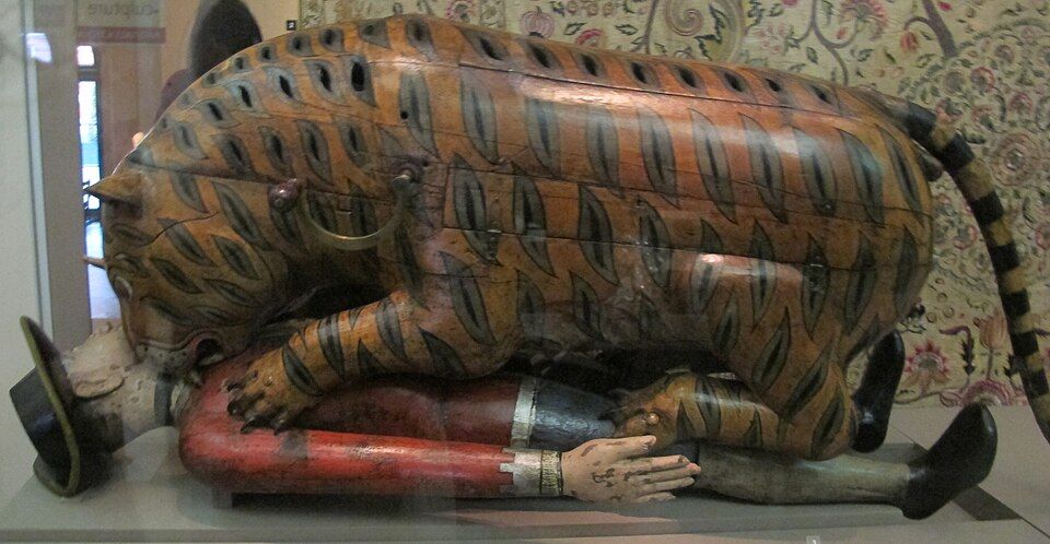 The wooden tiger and European figure in a museum case with a patterned textile behind them.