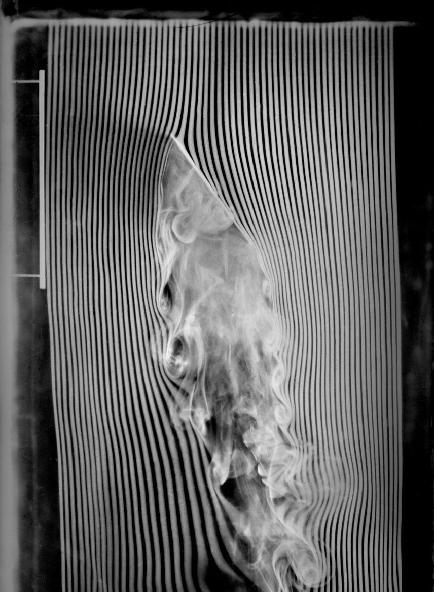 Bright threads of smoke bend around an inclined plate and break into curls in its wake, against a black background.