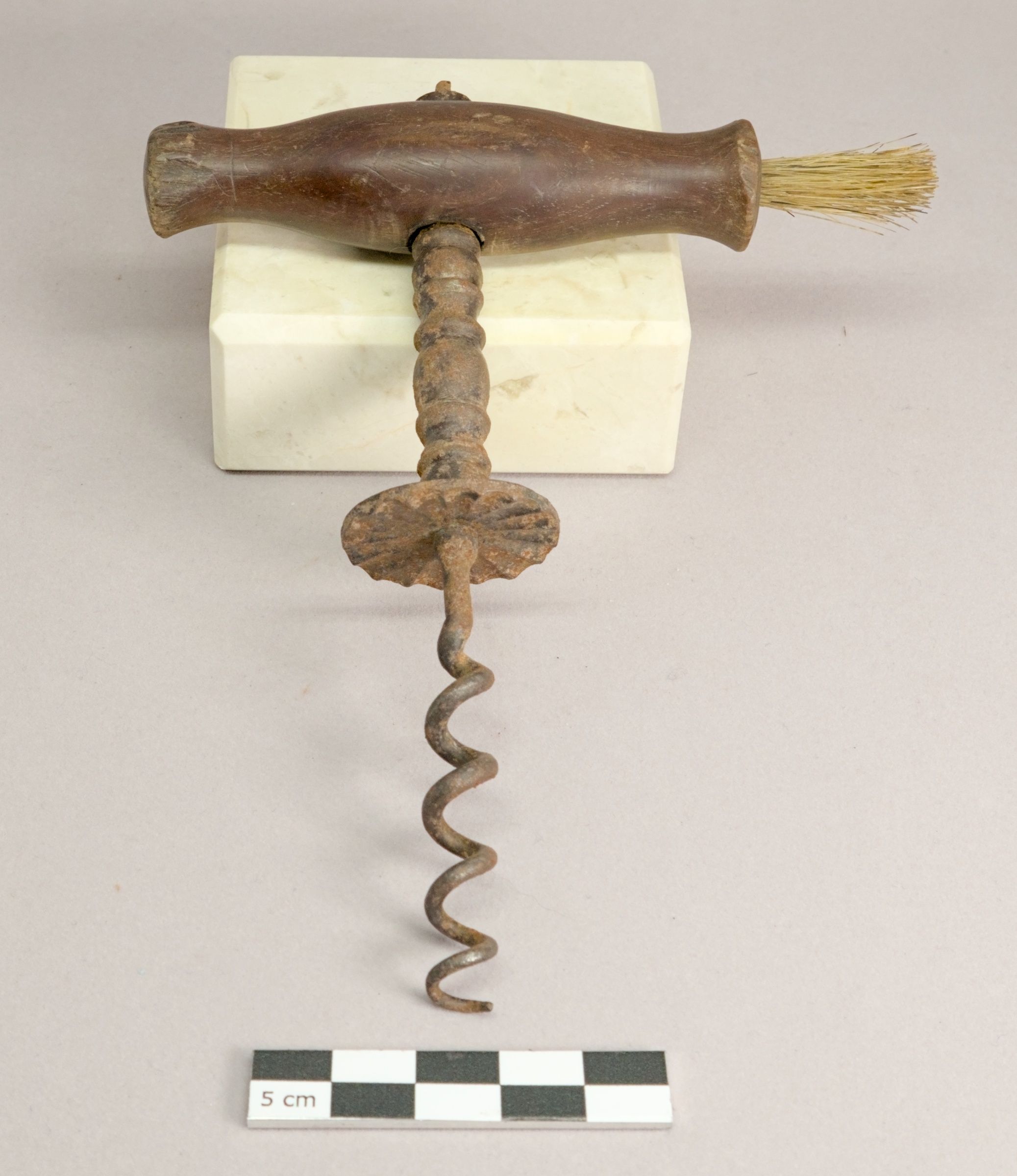 A wooden-handled corkscrew with a serrated disc above its open spiral and a brush at one end