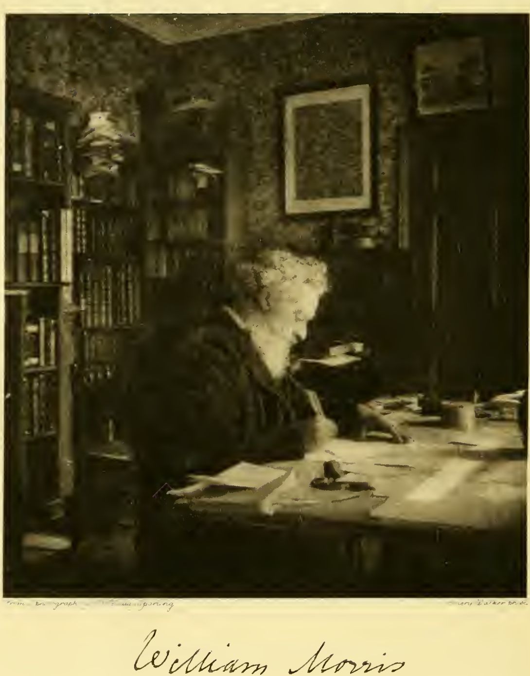 William Morris seated writing at a crowded desk, with bookshelves and framed pictures behind him