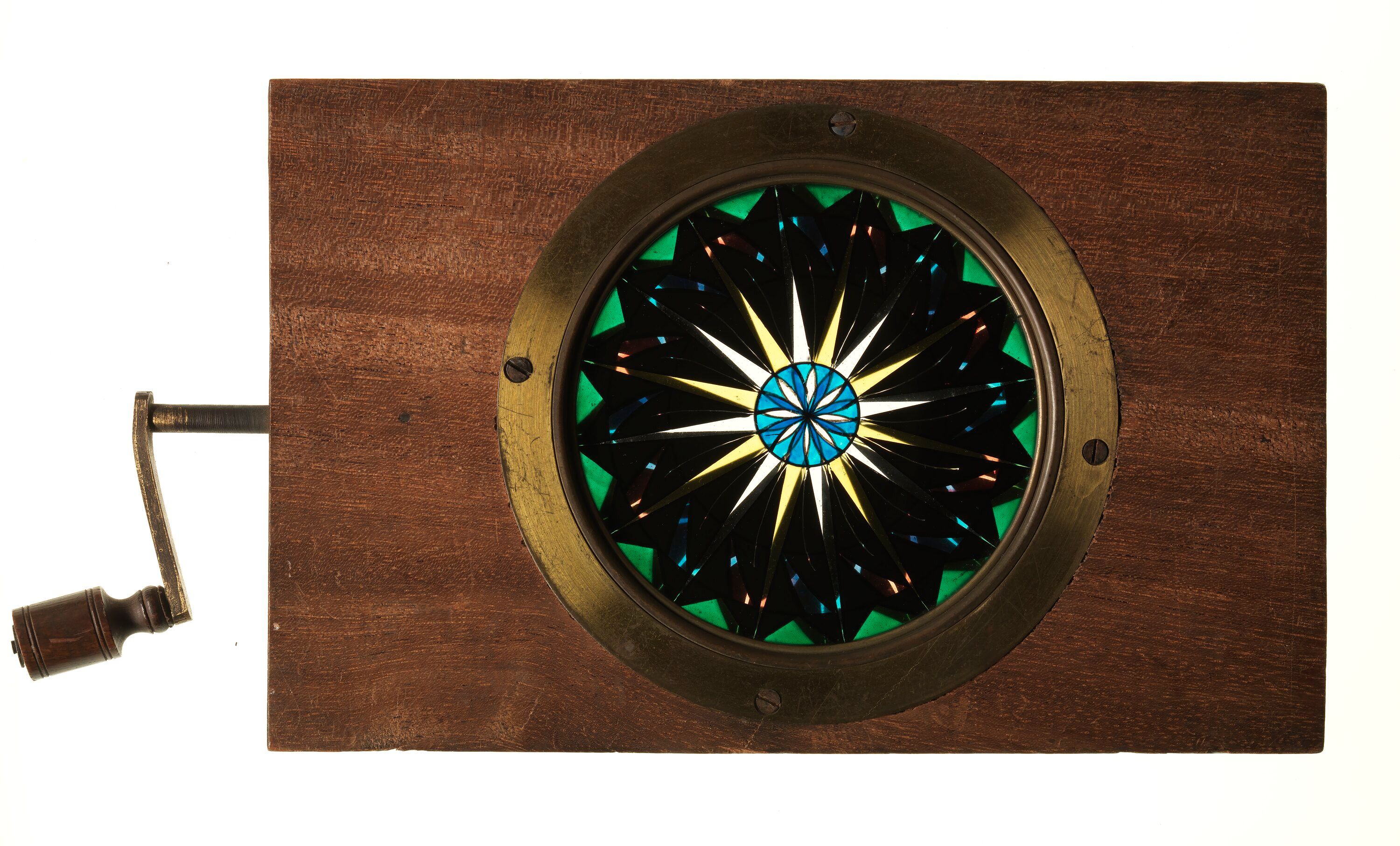 A circular blue, yellow and green pattern sits inside a brass ring in a wooden slide frame.