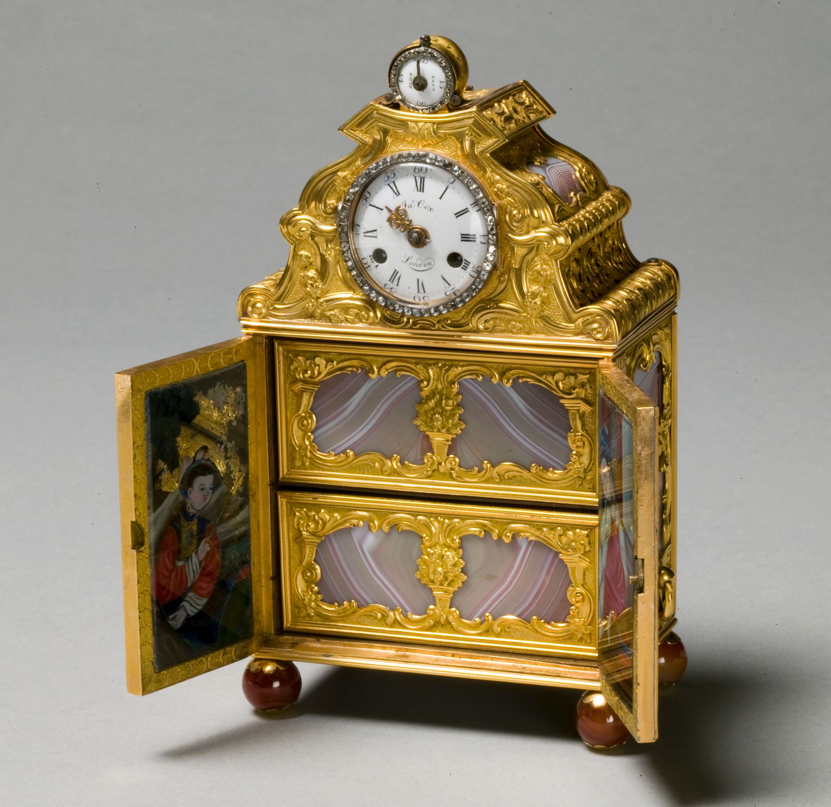 A small gilded cabinet with a clock dial, coloured panels and an elaborate crest.
