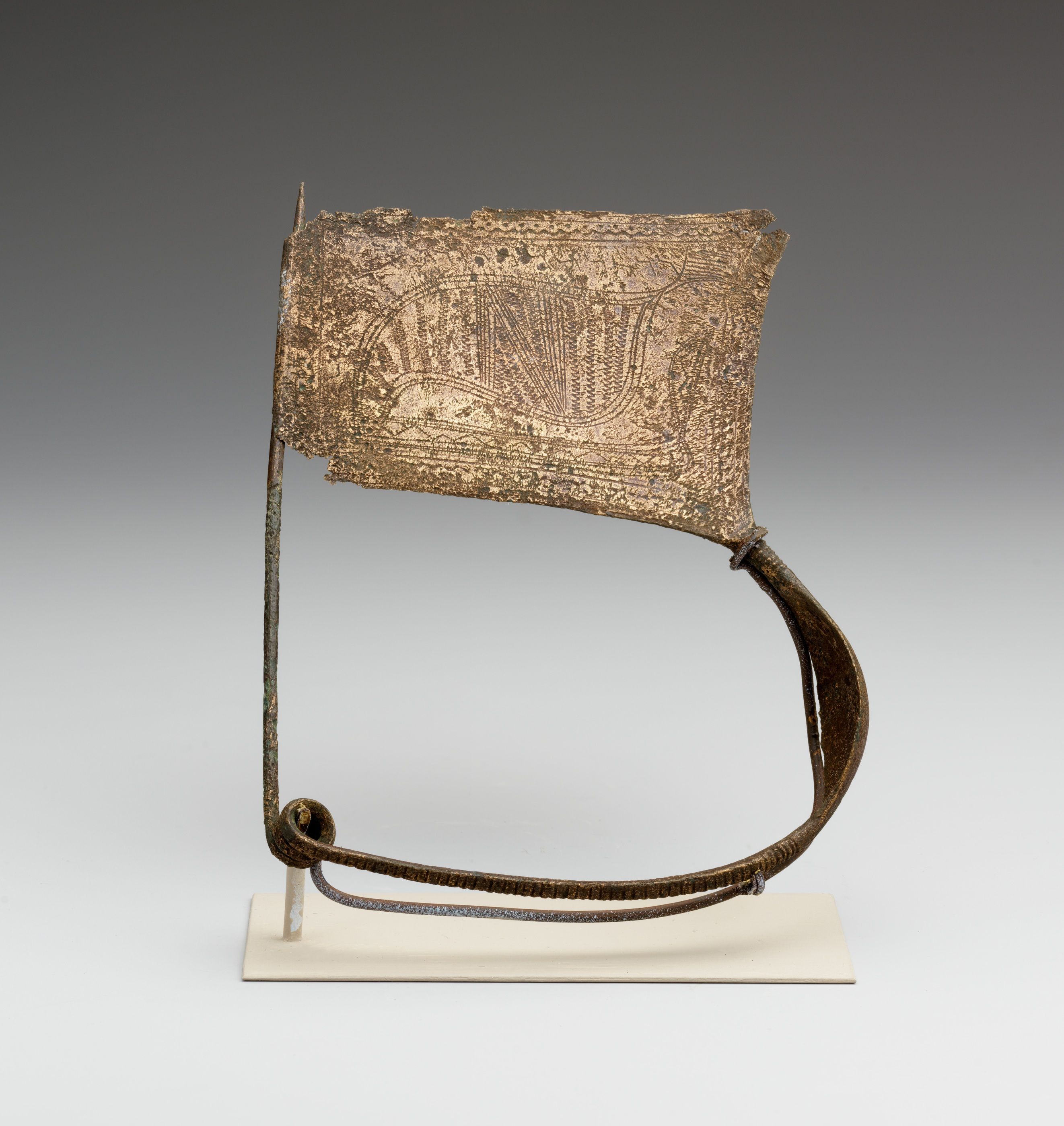 An ancient bronze clothing fastener with a coiled pin and a broad catch plate engraved with a bird