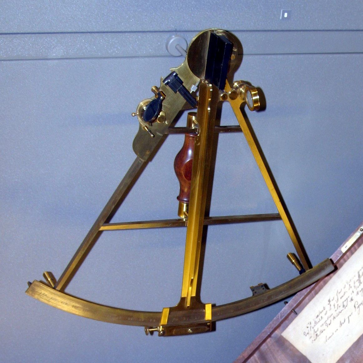 A brass sextant with mirrors and sights above an open frame, a wooden handle and a curved reading scale