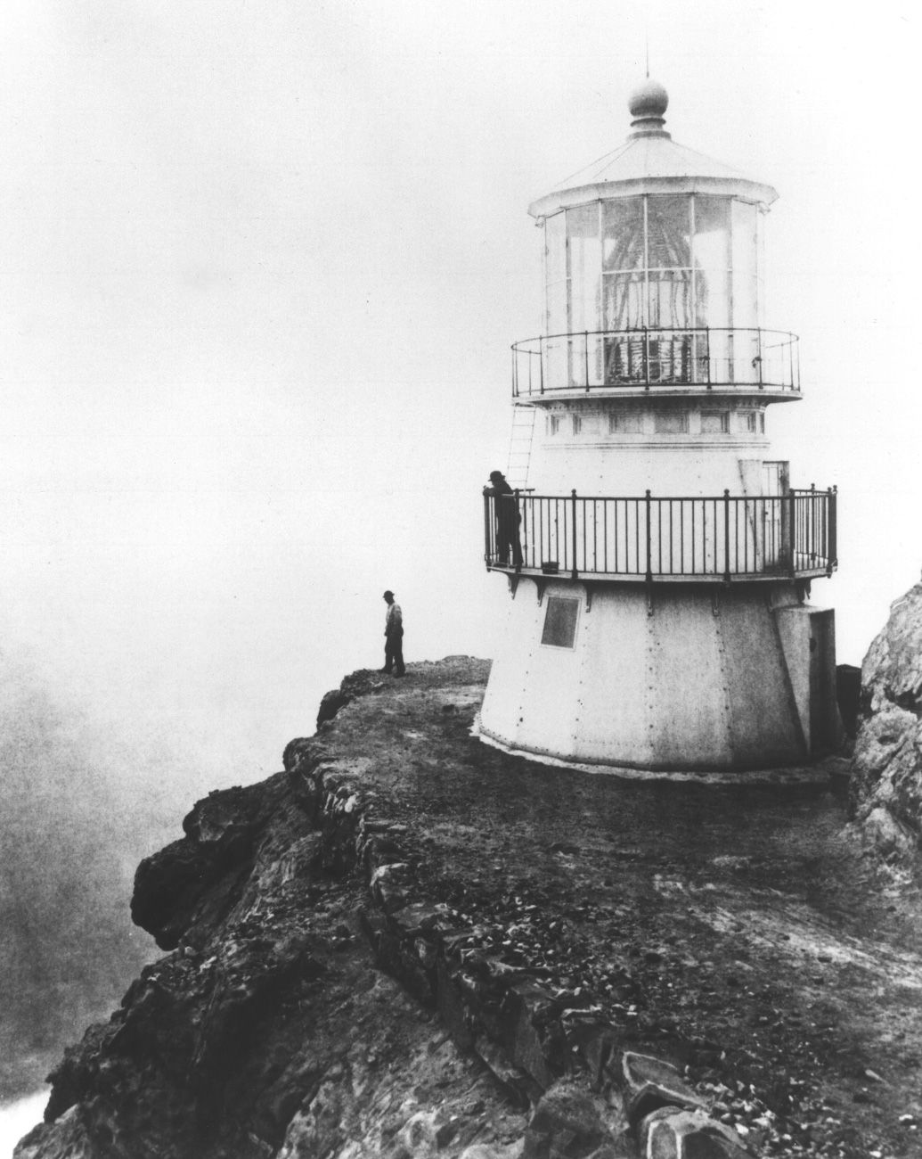 The newly built Point Reyes Lighthouse standing on a narrow rocky ledge above the sea, with two people beside it