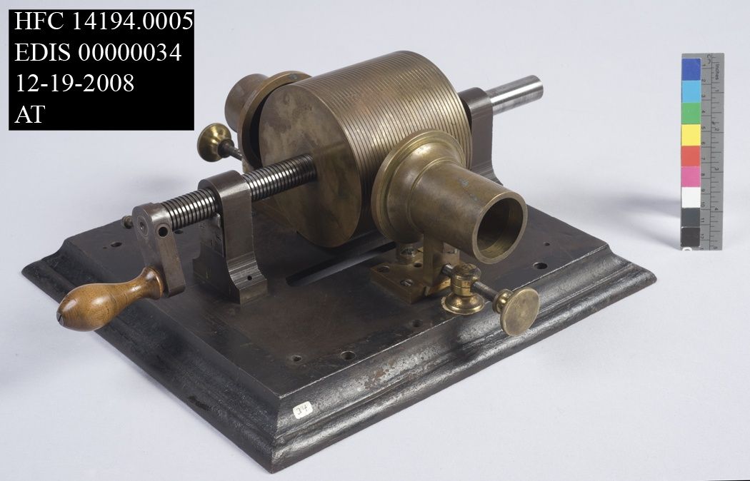 An early grooved brass cylinder phonograph on a wooden base, with a threaded shaft, hand crank and mouthpiece