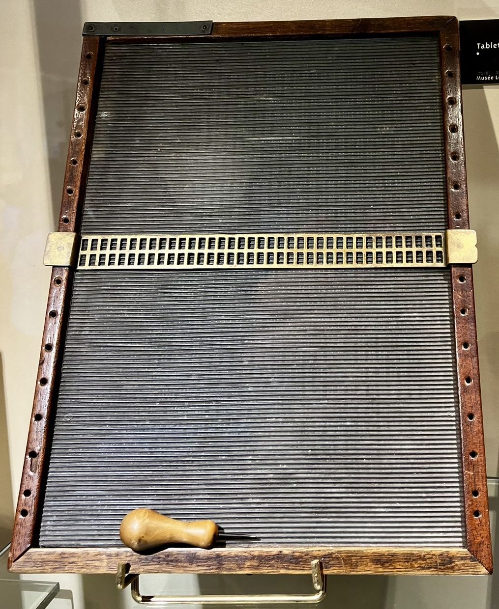 A wooden writing frame with a grooved bed, a two-line brass guide and a yellow-handled stylus displayed below it
