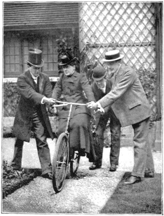 Willard seated on a low bicycle while three men steady it
