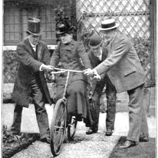 Willard seated on a low bicycle while three men steady it