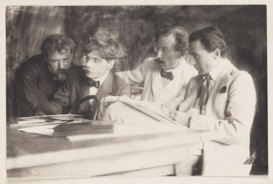 Four photographers lean towards prints spread on a table in a softly rendered black-and-white photograph.