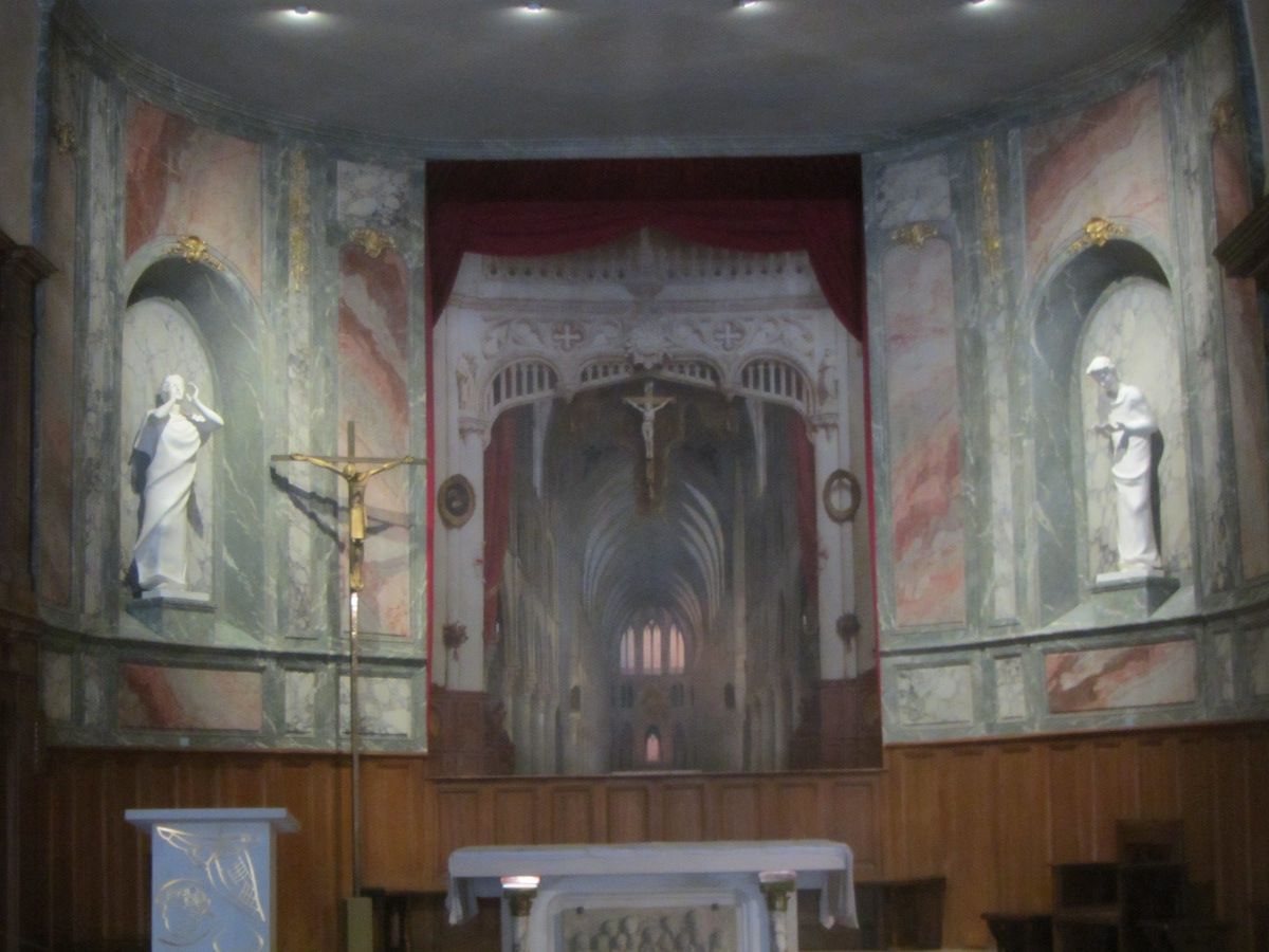 A painted Gothic nave recedes behind the altar of a small church, framed by real statues, marbled walls and a red curtain.