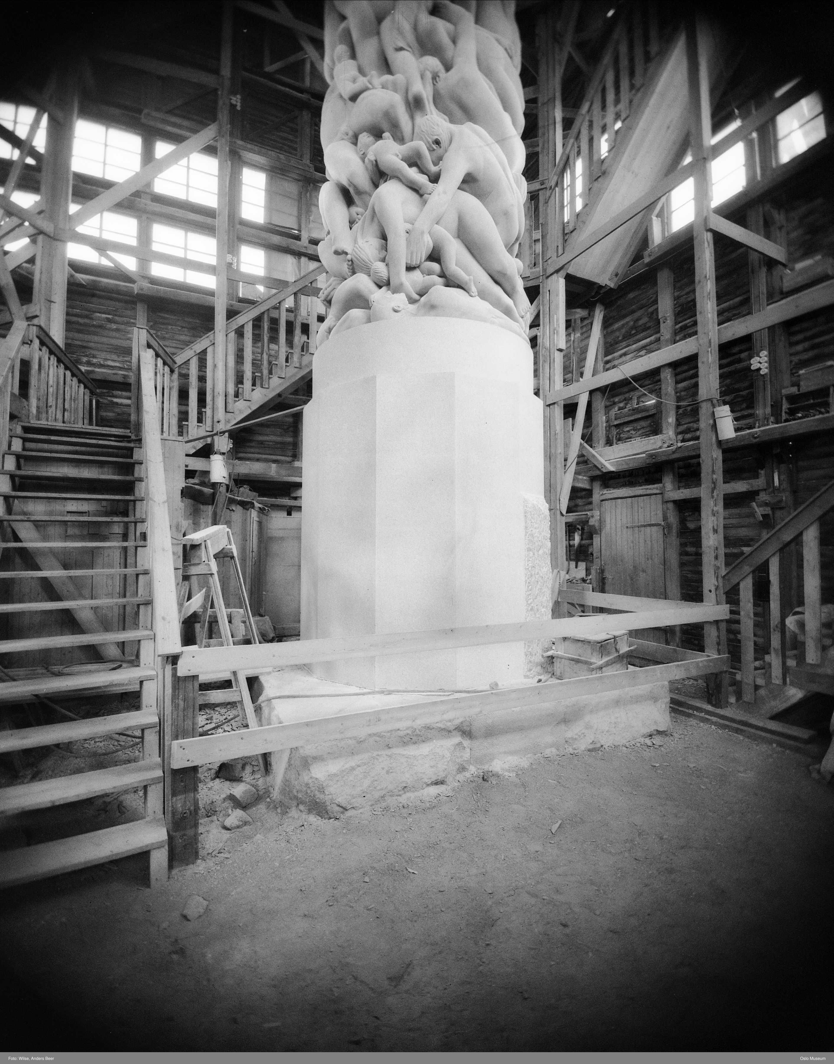 Upper human figures already carved, above a squared unfinished section of granite, inside timber scaffolding.