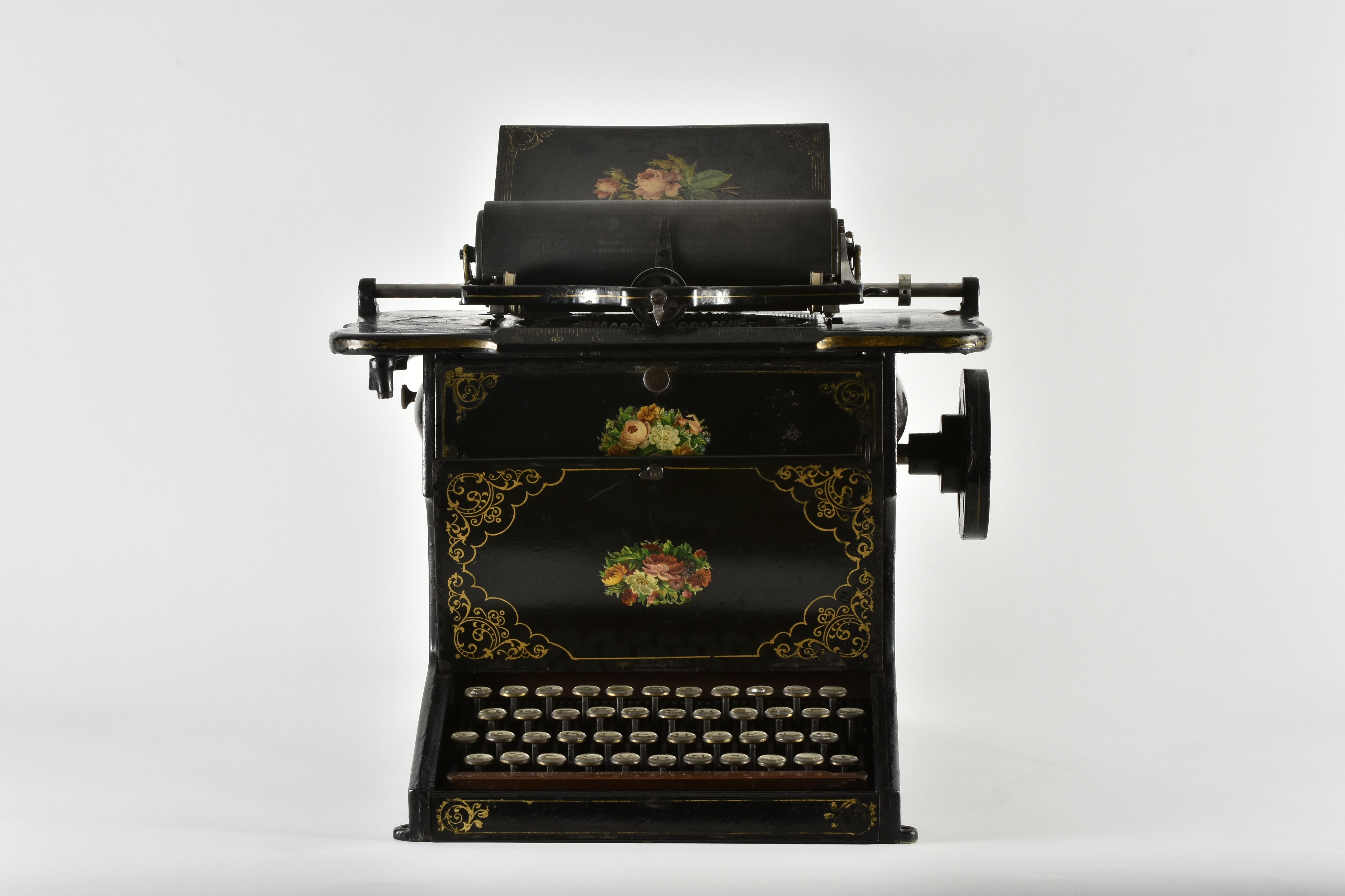 A black Sholes & Glidden typewriter with gold floral decoration, round keys and a paper roller above the mechanism