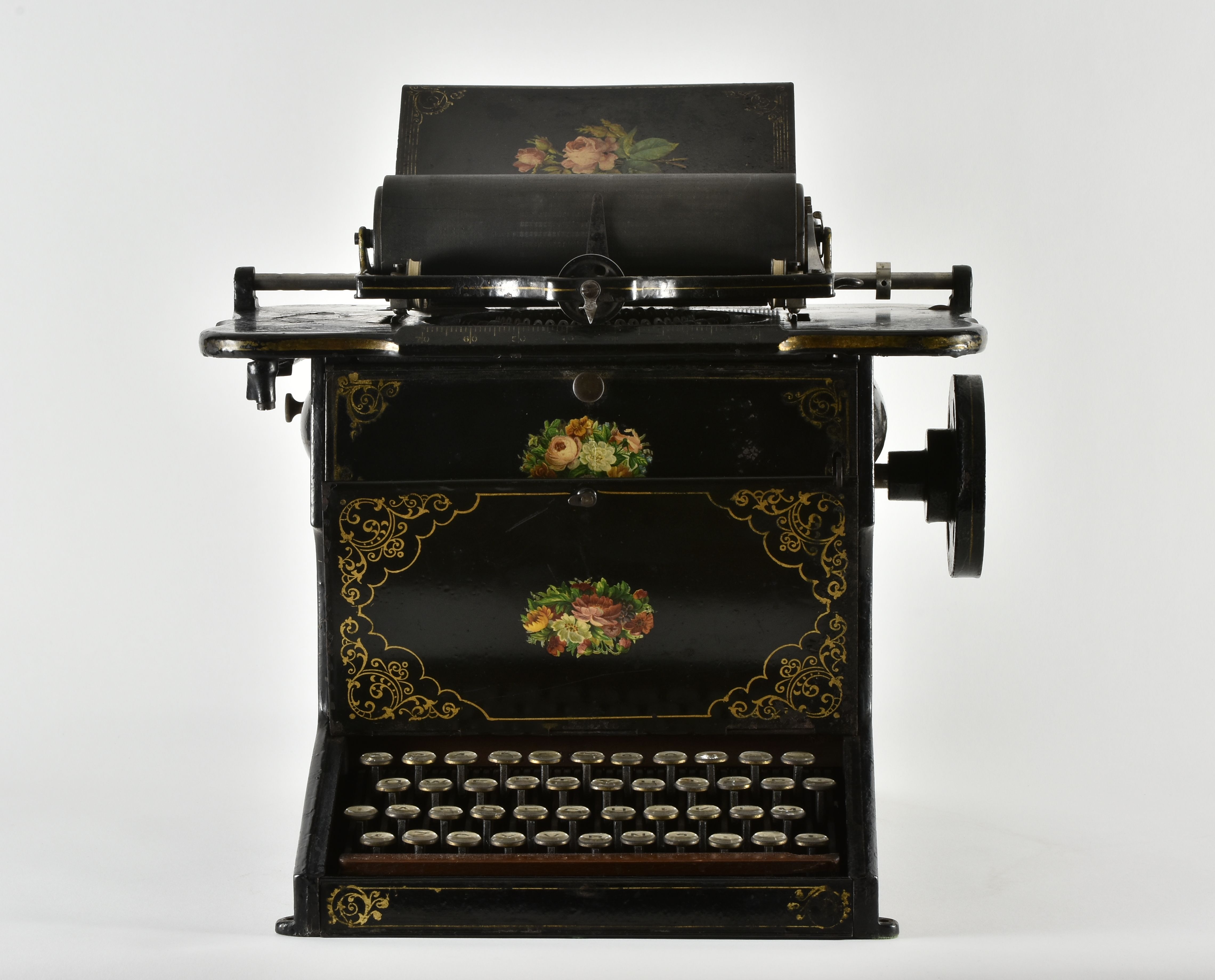 A black Sholes & Glidden typewriter with gold floral decoration, round keys and a paper roller above the mechanism