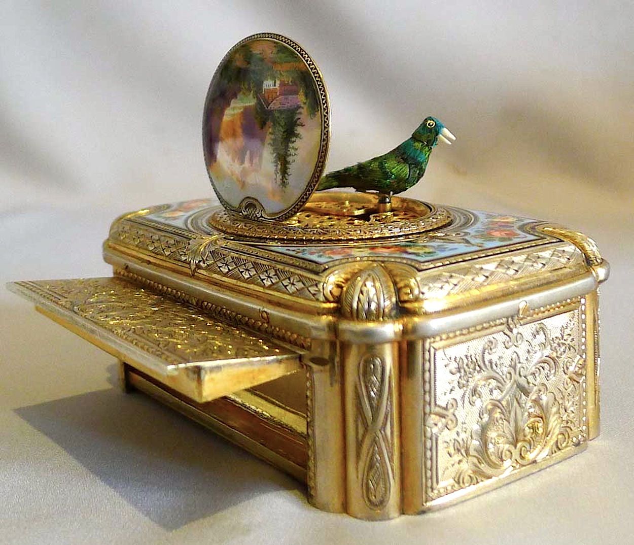 A green feathered mechanical bird stands above an open silver-gilt box, beside its raised enamel lid and open tobacco compartment