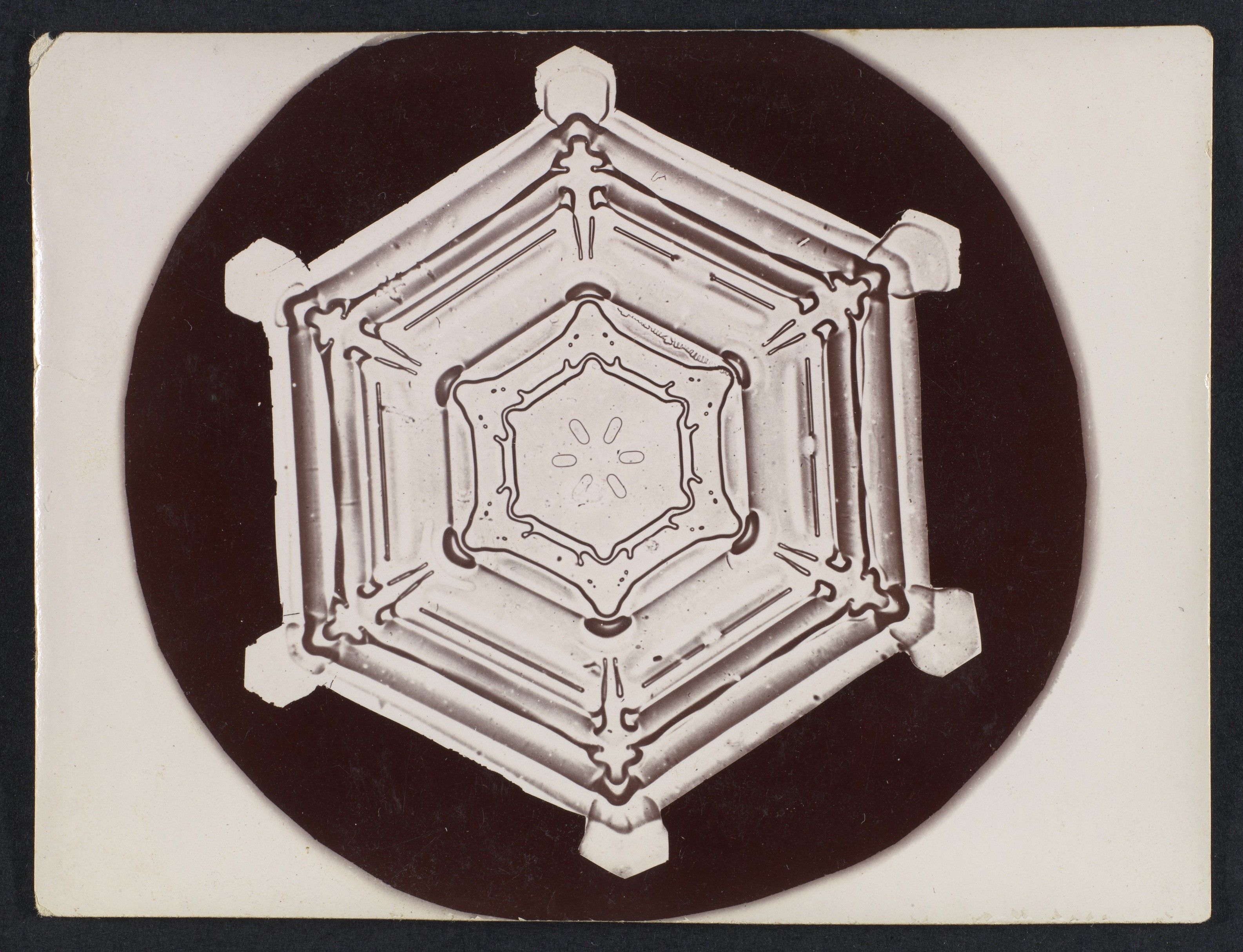 A compact hexagonal snow crystal with layered internal markings, surrounded by a dark circular field.