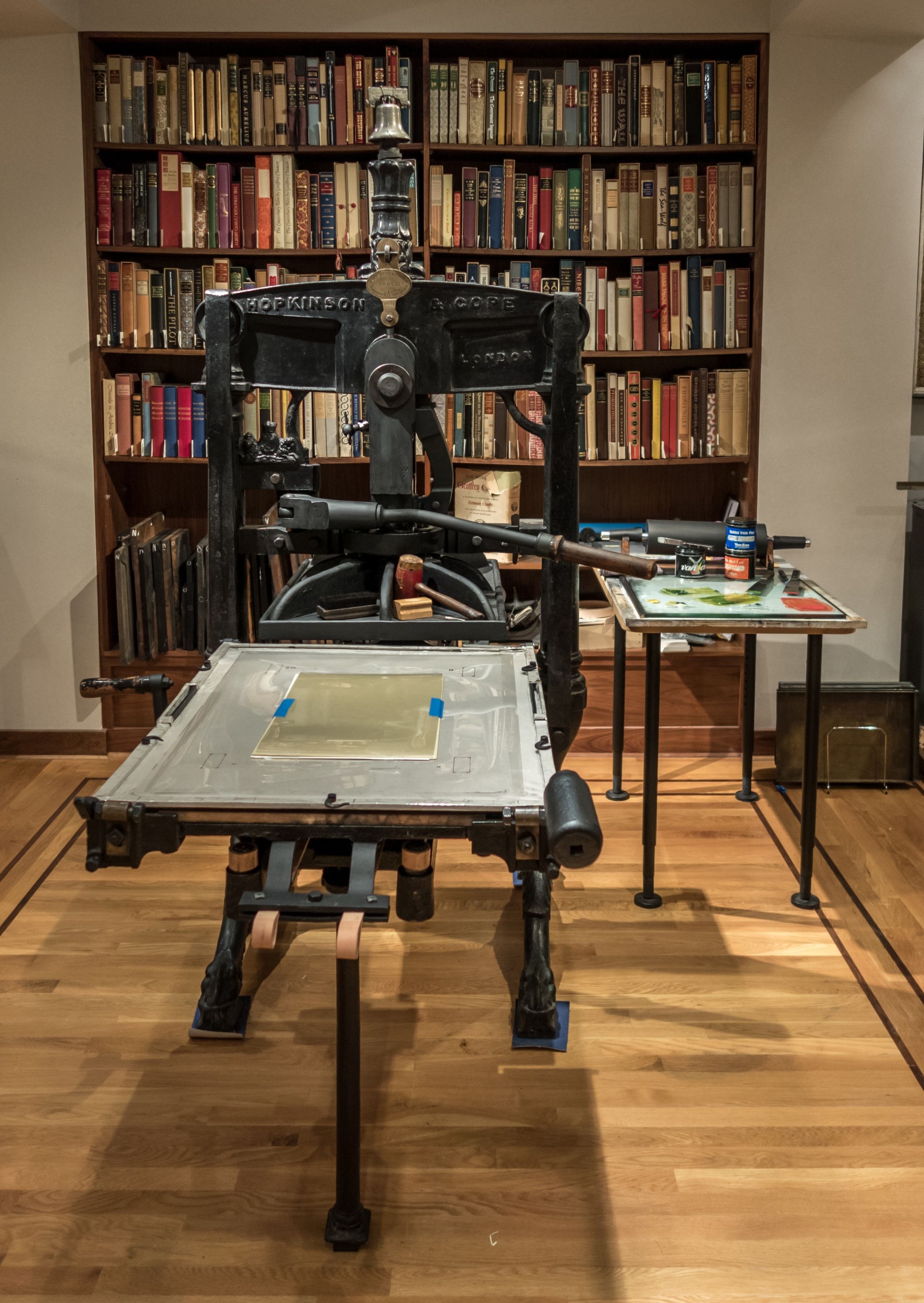 A tall iron Albion hand press with a long handle, raised paper frame and a printing bed in a modern library