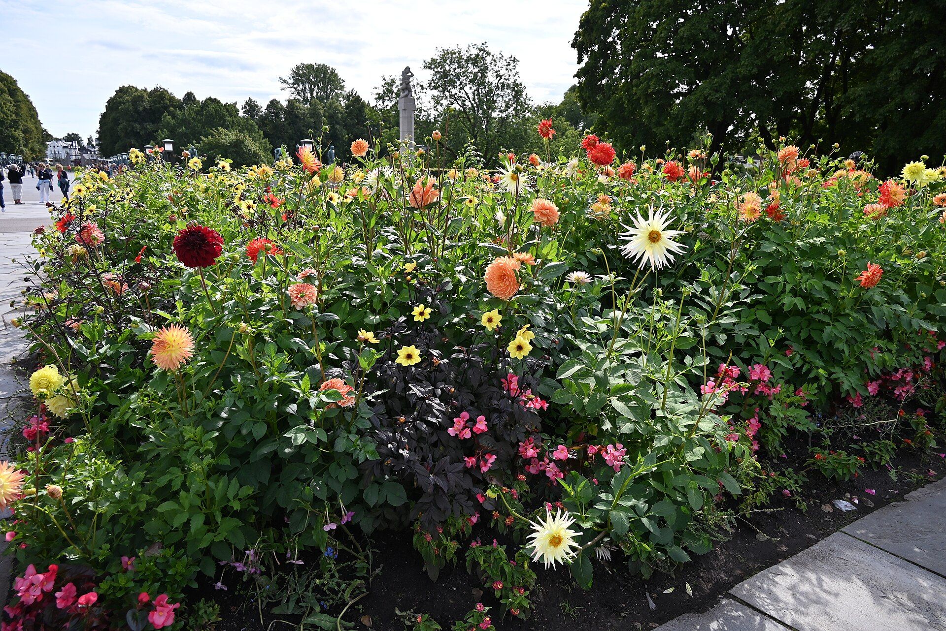Pink and red flowers beneath mature trees in Frogner Park, with a bronze sculpture beyond.