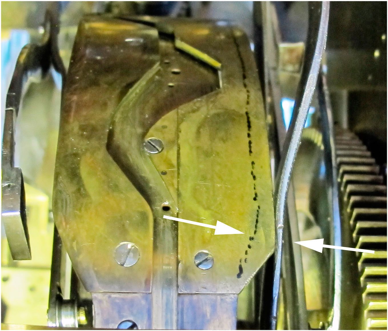 A close view of a brass cam track beside a narrow iron lever, with white arrows marking their contact.