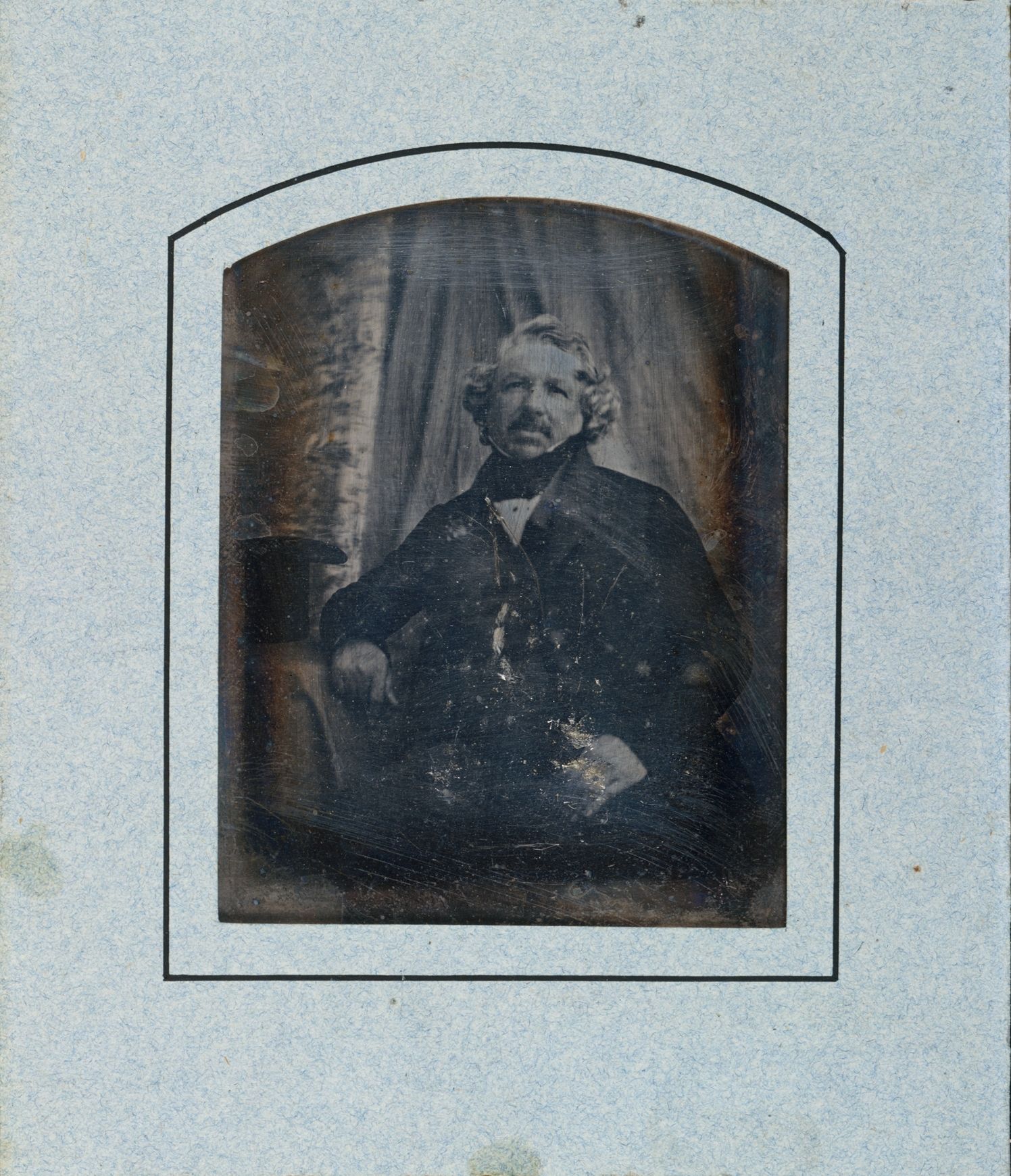 Louis Daguerre sits with one arm resting on a table, looking towards the camera; the surviving photographic plate bears scratches and damage.