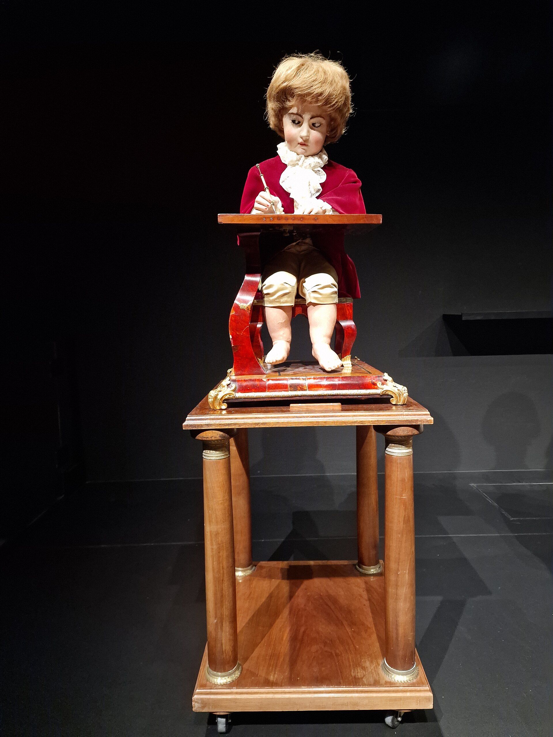 The blond-haired Draughtsman seated at a small drawing table.